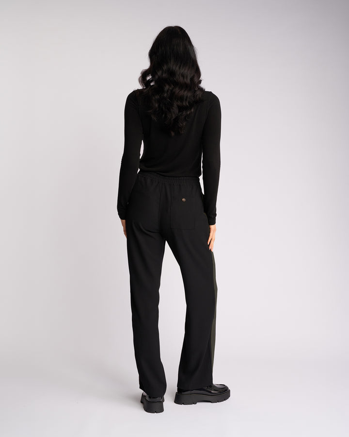 A person with long, wavy dark hair, dressed in a black long-sleeve top and the Knewe Label Bowie Pant - Black/Moss, featuring a pull-on style and elasticated waistband, stands facing away from the camera against a plain white background.