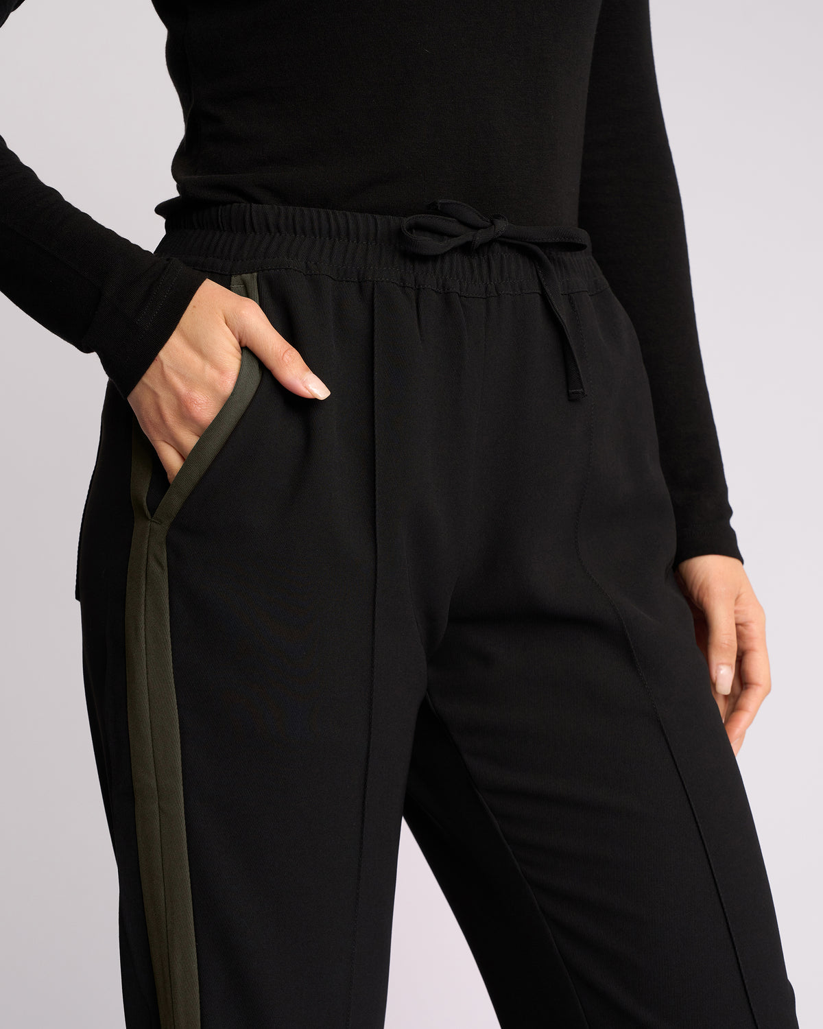A person wearing the Knewe Label Bowie Pant in Black/Moss, featuring straight legs, an elastic waistband, and an olive green side stripe, pairs it with a black long-sleeve top; one hand is in their pocket and the other at their side.