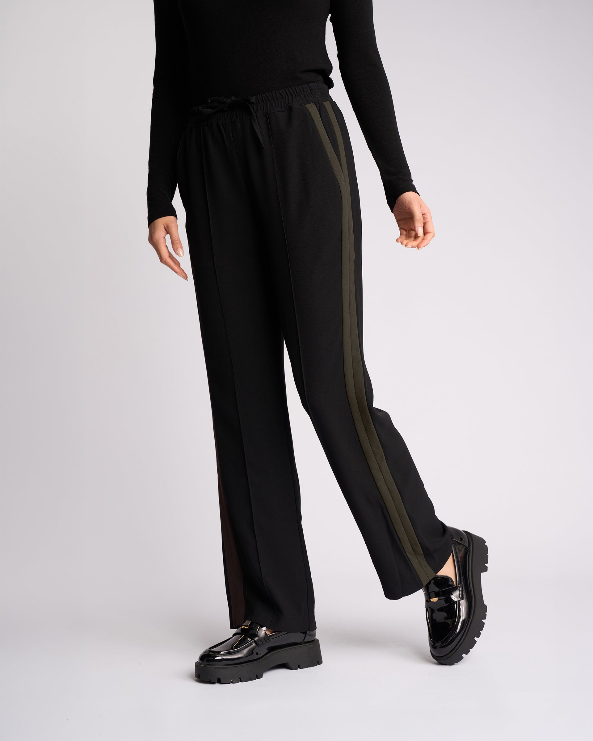 A person stands against a plain light background wearing the Knewe Label Bowie Pant - Black/Moss, featuring black pull-on wide-leg pants with olive green side stripes. They pair it with a black long-sleeve top and shiny black loafers; their face is out of frame.