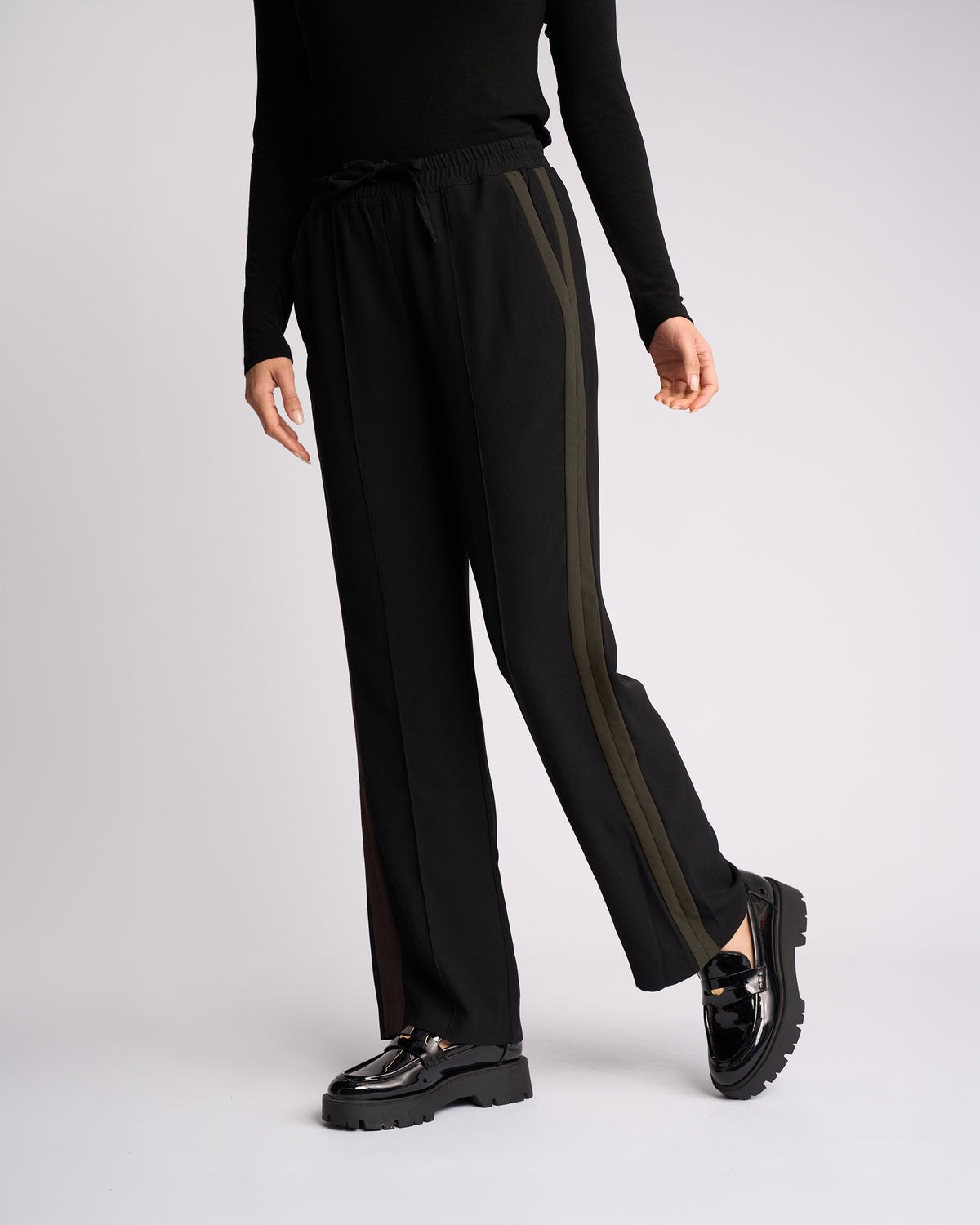 A person stands against a plain light background wearing the Knewe Label Bowie Pant - Black/Moss, featuring black pull-on wide-leg pants with olive green side stripes. They pair it with a black long-sleeve top and shiny black loafers; their face is out of frame.