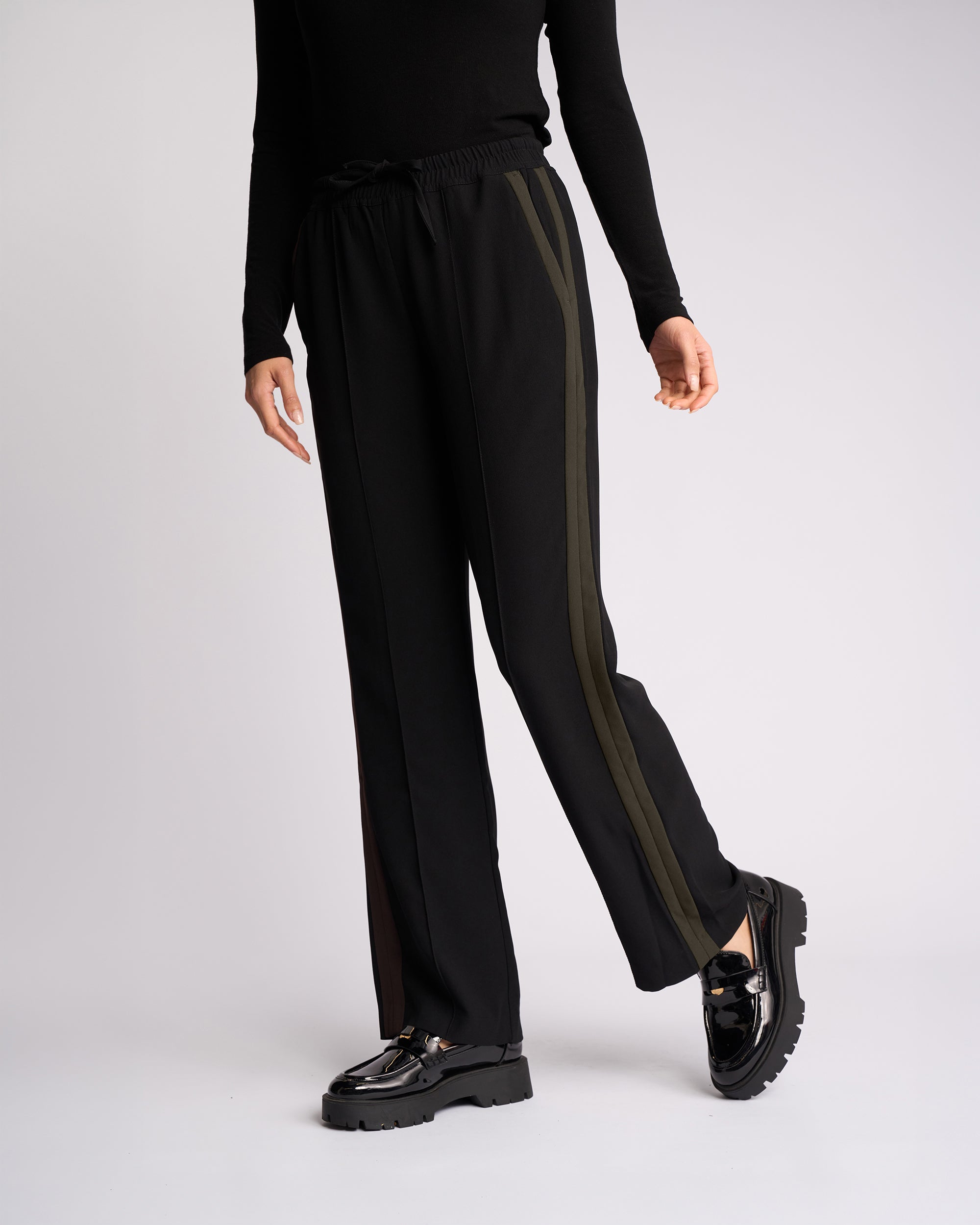 A person stands against a plain light background wearing the Knewe Label Bowie Pant - Black/Moss, featuring black pull-on wide-leg pants with olive green side stripes. They pair it with a black long-sleeve top and shiny black loafers; their face is out of frame.