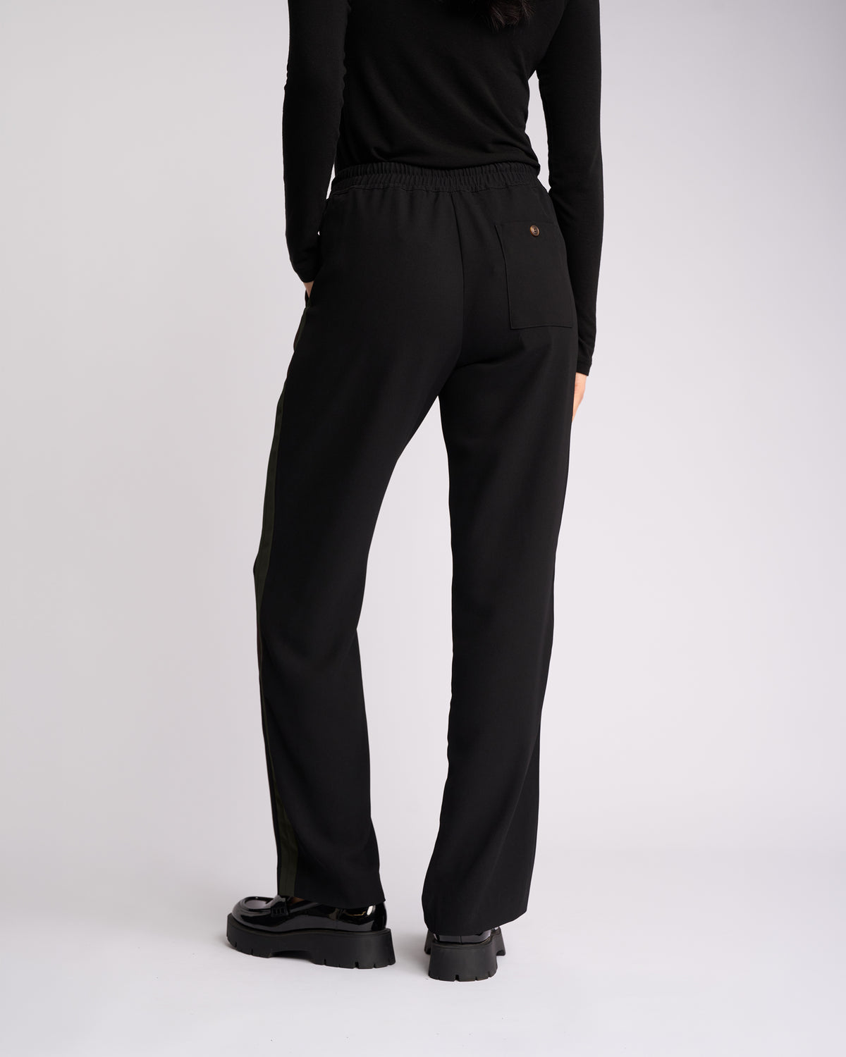 Person standing with their back to the camera, wearing a black long-sleeve top, Knewe Label Bowie Pant in Black/Moss—featuring a straight-leg pull-on design with an elasticated waistband—and black chunky shoes, against a plain light background.