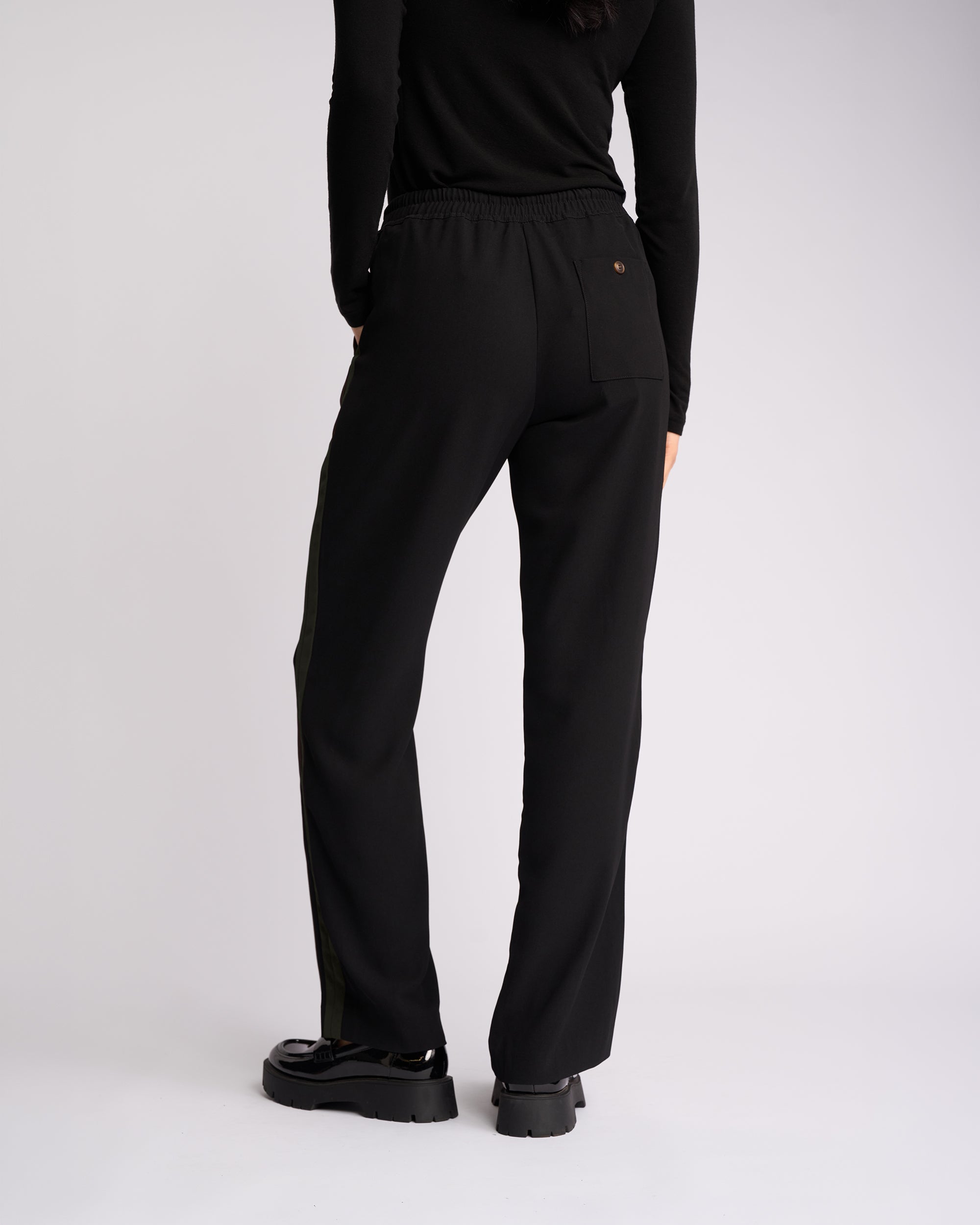 Person standing with their back to the camera, wearing a black long-sleeve top, Knewe Label Bowie Pant in Black/Moss—featuring a straight-leg pull-on design with an elasticated waistband—and black chunky shoes, against a plain light background.