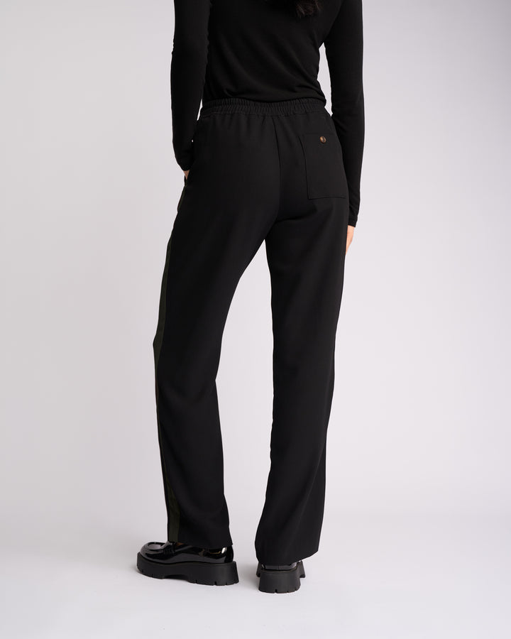 Person standing with their back to the camera, wearing a black long-sleeve top, Knewe Label Bowie Pant in Black/Moss—featuring a straight-leg pull-on design with an elasticated waistband—and black chunky shoes, against a plain light background.