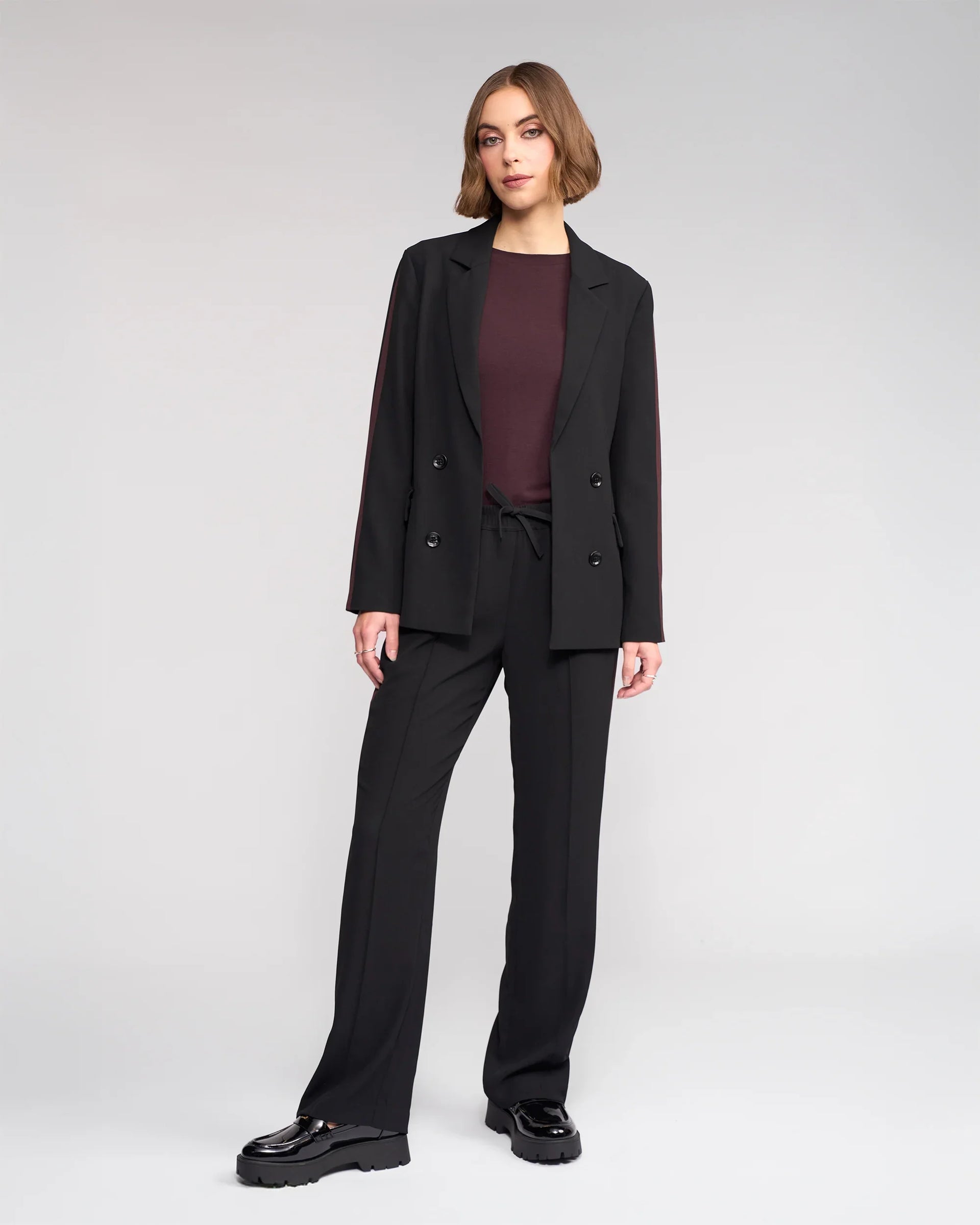 A woman with short brown hair stands against a light gray background in the Knewe Label Bowie Pant - Black/Raisin, featuring a straight-leg cut and elasticated waistband, paired with a black blazer, burgundy top, and black platform shoes.