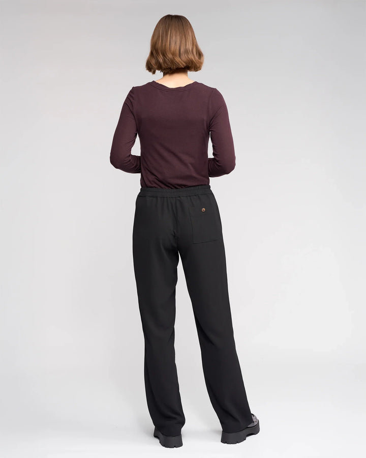 A person with short brown hair, wearing a long-sleeve dark maroon top and Knewe Label’s Bowie Pant in Black/Raisin, featuring an elasticated waistband, stands facing away from the camera against a light gray background.