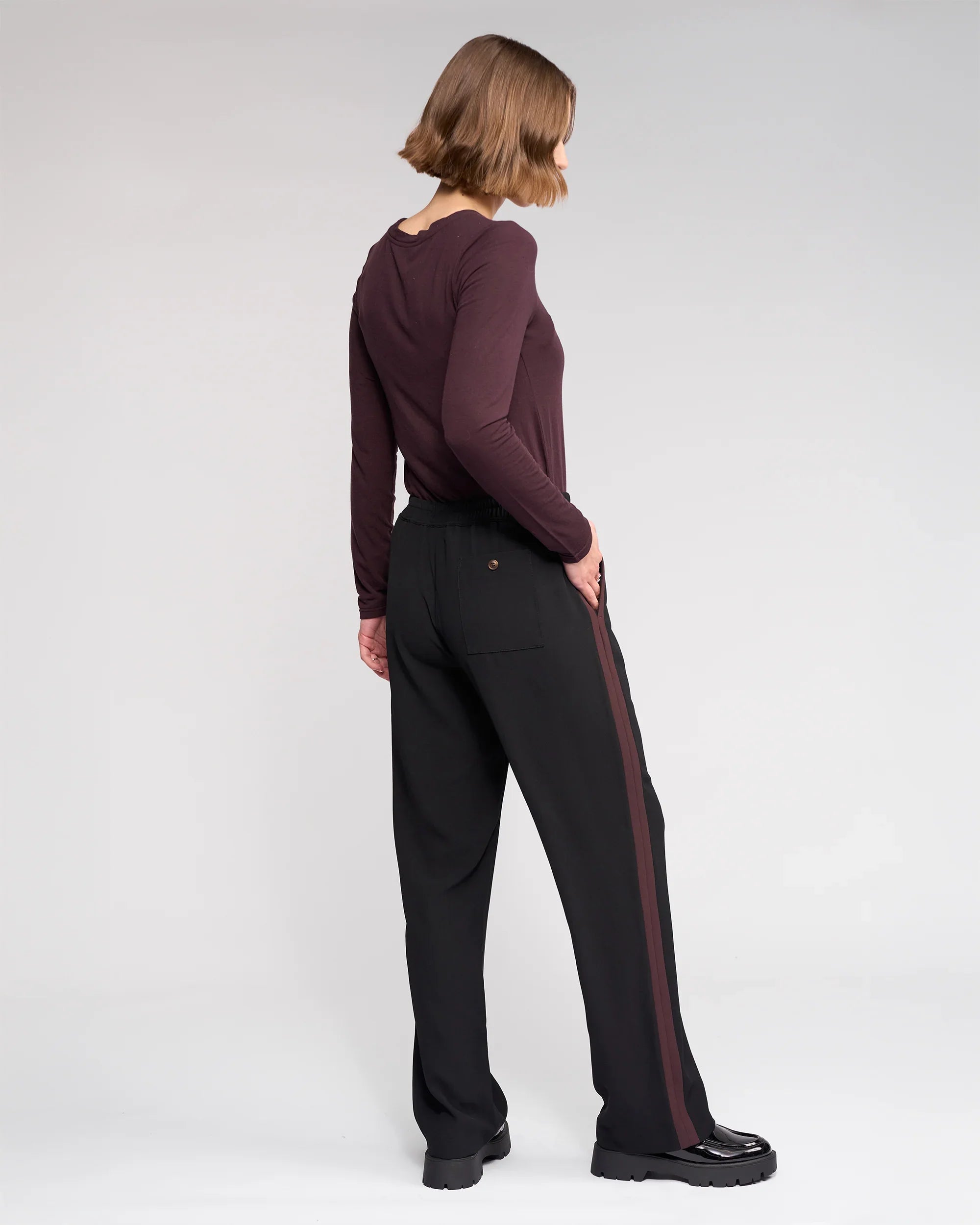 A person with chin-length brown hair, facing sideways, wears a maroon top and the Knewe Label Bowie Pant - Black/Raisin, featuring a brown side stripe and elasticated waistband, plus black shoes, set against a plain light gray background.