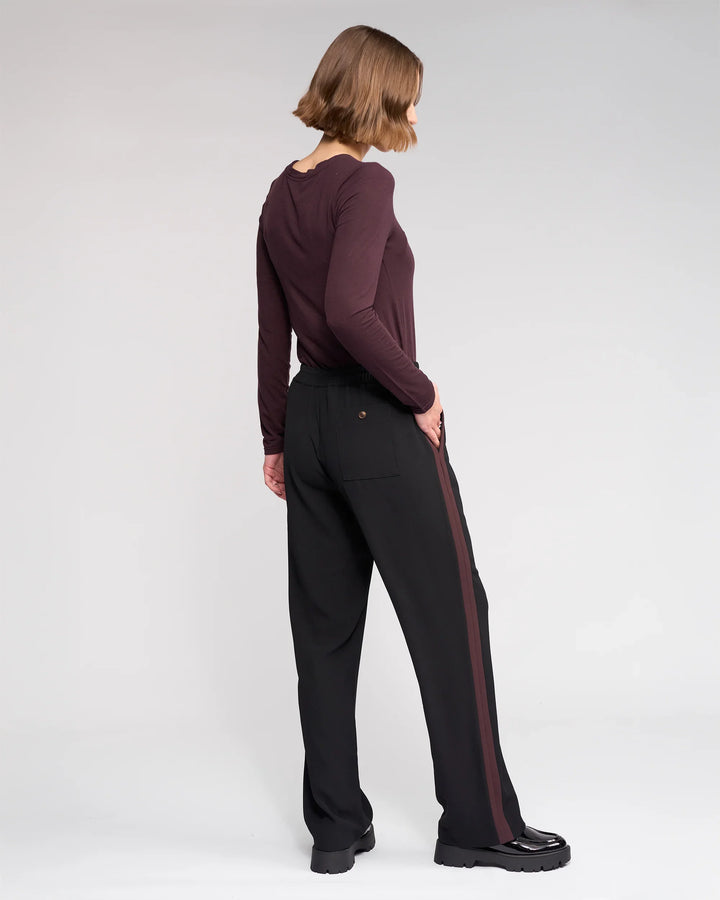 A person with chin-length brown hair, facing sideways, wears a maroon top and the Knewe Label Bowie Pant - Black/Raisin, featuring a brown side stripe and elasticated waistband, plus black shoes, set against a plain light gray background.
