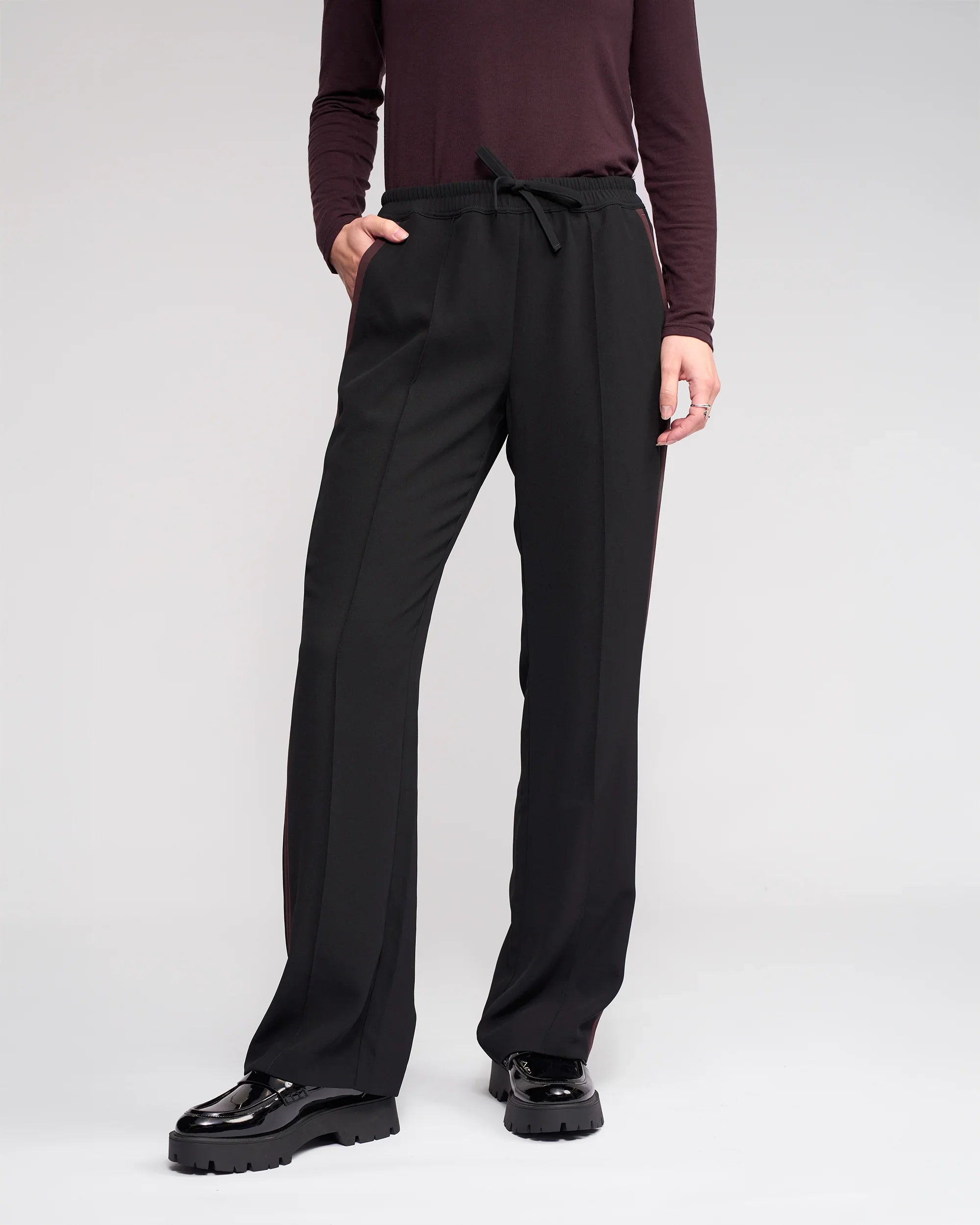 Wearing the Knewe Label Bowie Pant in Black/Raisin and a long-sleeve burgundy top, a person stands with one hand in their pocket against a plain background, paired with black chunky shoes.