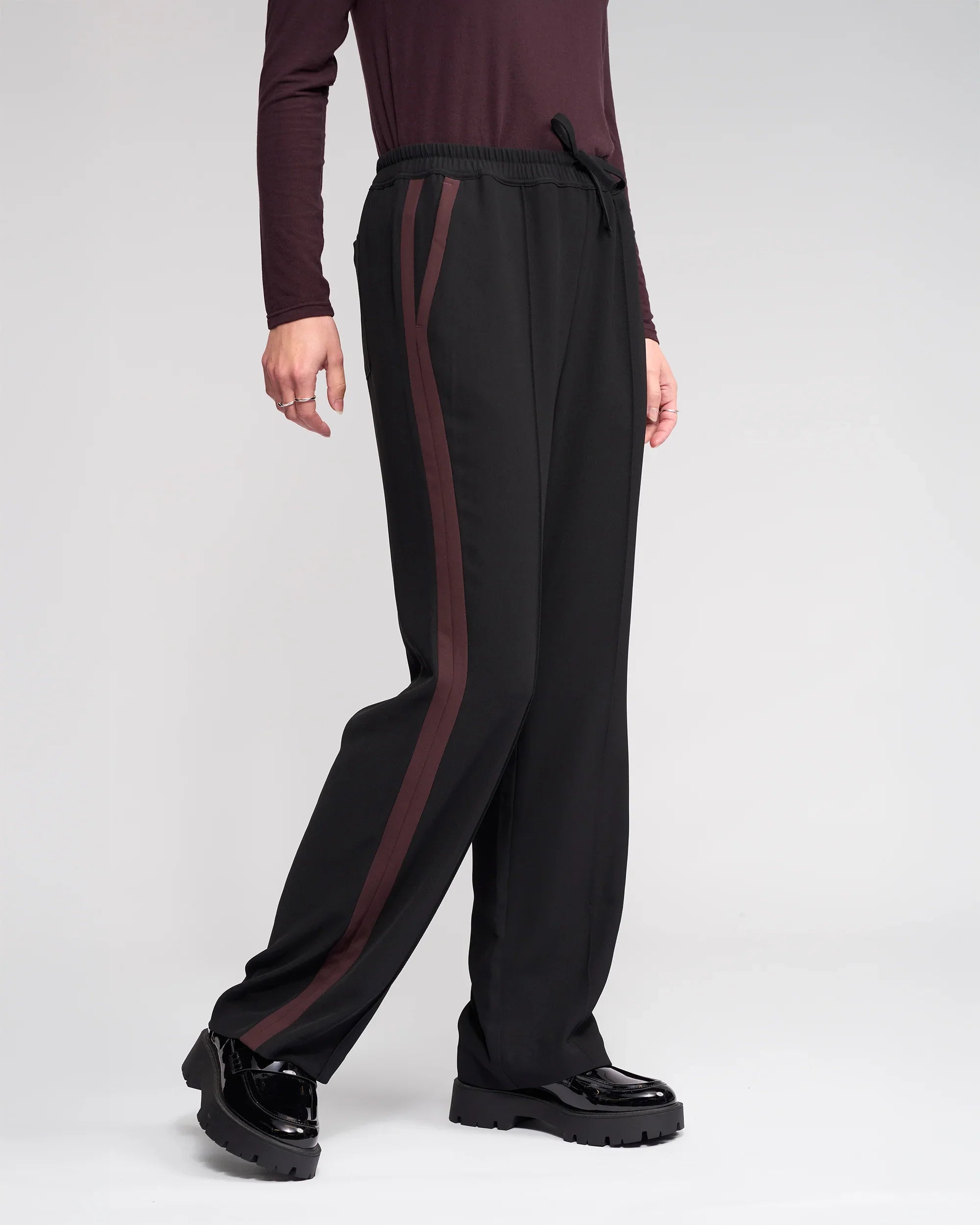 Person wearing Knewe Label’s Bowie Pant in Black/Raisin with maroon side stripes and an elastic waistband, paired with a matching maroon top and black shiny shoes, standing against a plain light background for relaxed comfort.