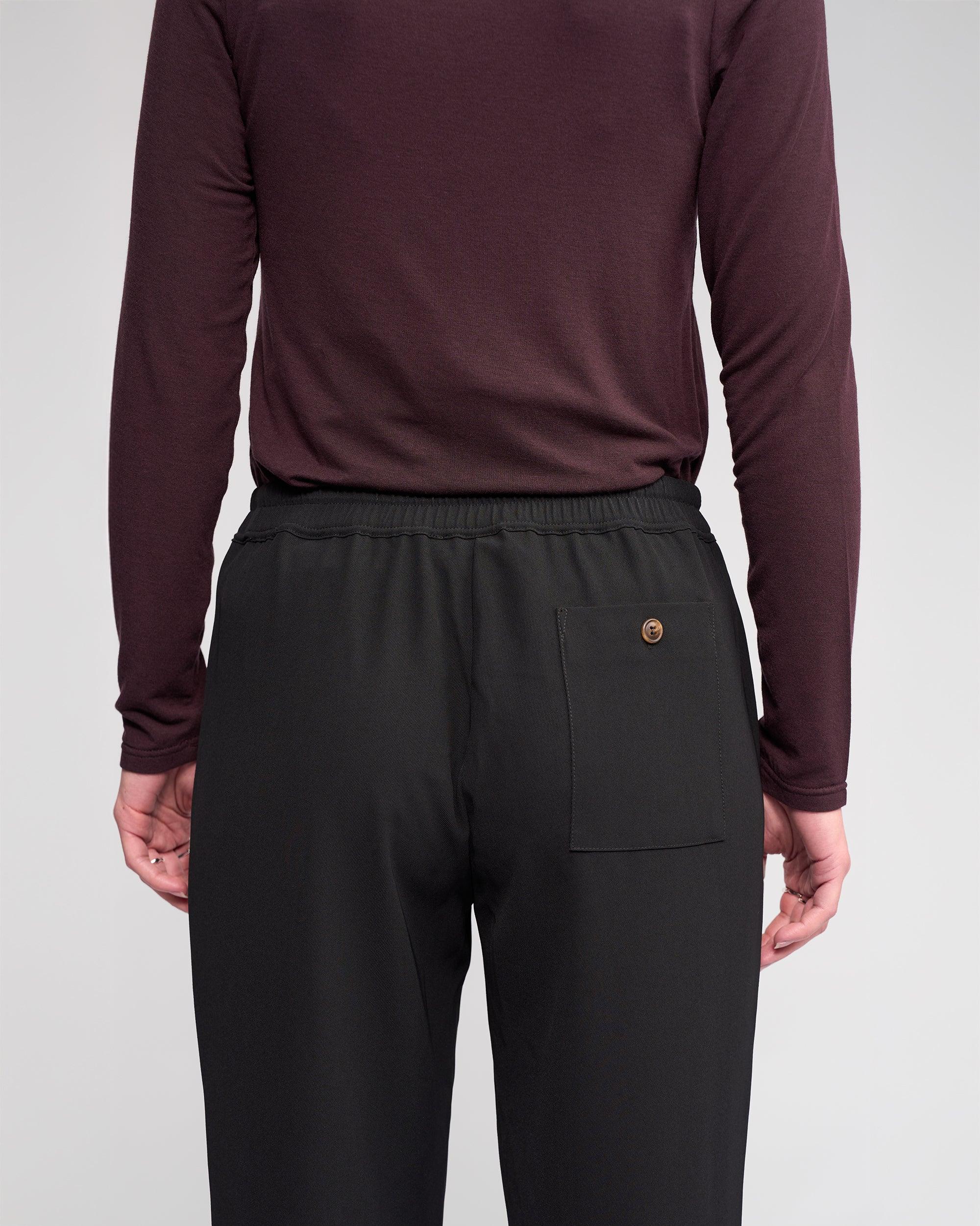 A person faces away from the camera, wearing Knewe Label’s Bowie Pant in Black/Raisin—straight-leg trousers with an elasticated waistband and a single right back pocket for relaxed comfort—and a long-sleeved burgundy shirt.