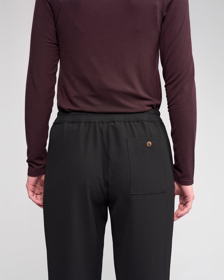 A person faces away from the camera, wearing Knewe Label’s Bowie Pant in Black/Raisin—straight-leg trousers with an elasticated waistband and a single right back pocket for relaxed comfort—and a long-sleeved burgundy shirt.