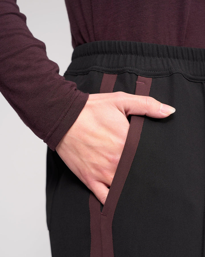 A close-up of a person wearing Knewe Label's Bowie Pant in Black/Raisin, featuring straight legs, maroon stripes, an elasticated waistband, and their right hand tucked into the pocket.