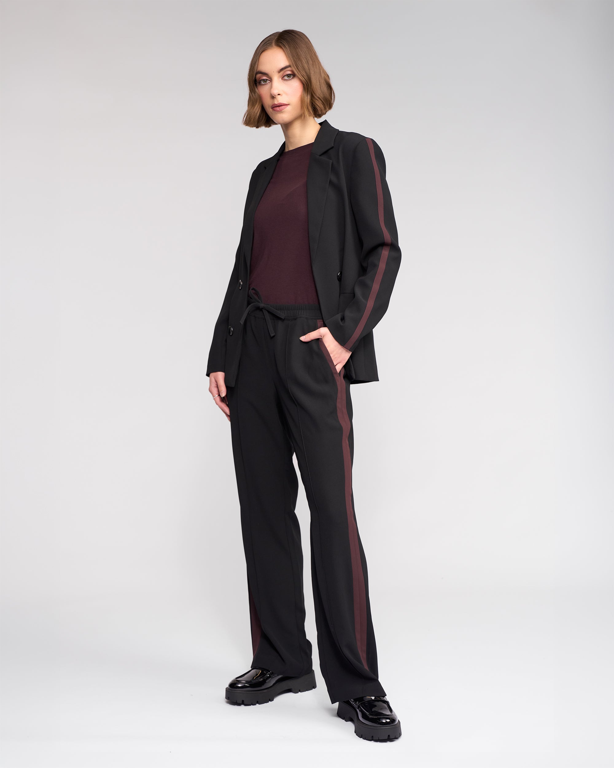 A woman with short brown hair wears the Knewe Label Bowie Pant in Black/Raisin, styled with a maroon top and chunky black shoes, paired with a black blazer, standing against a plain light background with one hand in her pocket.