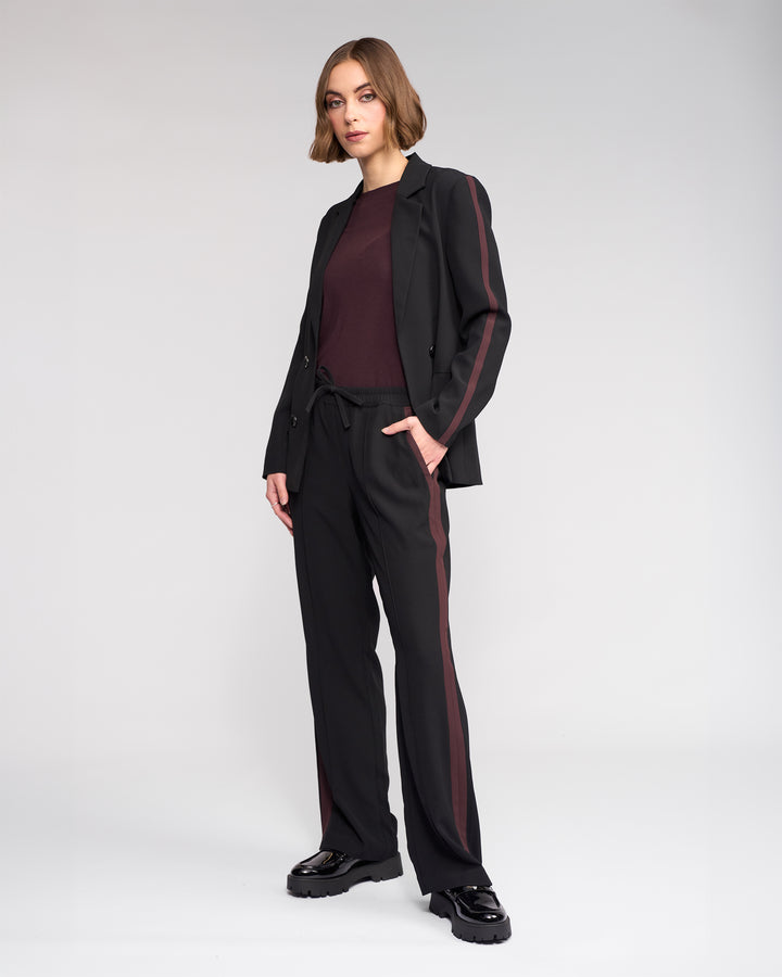 A woman with short brown hair wears the Knewe Label Bowie Pant in Black/Raisin, styled with a maroon top and chunky black shoes, paired with a black blazer, standing against a plain light background with one hand in her pocket.