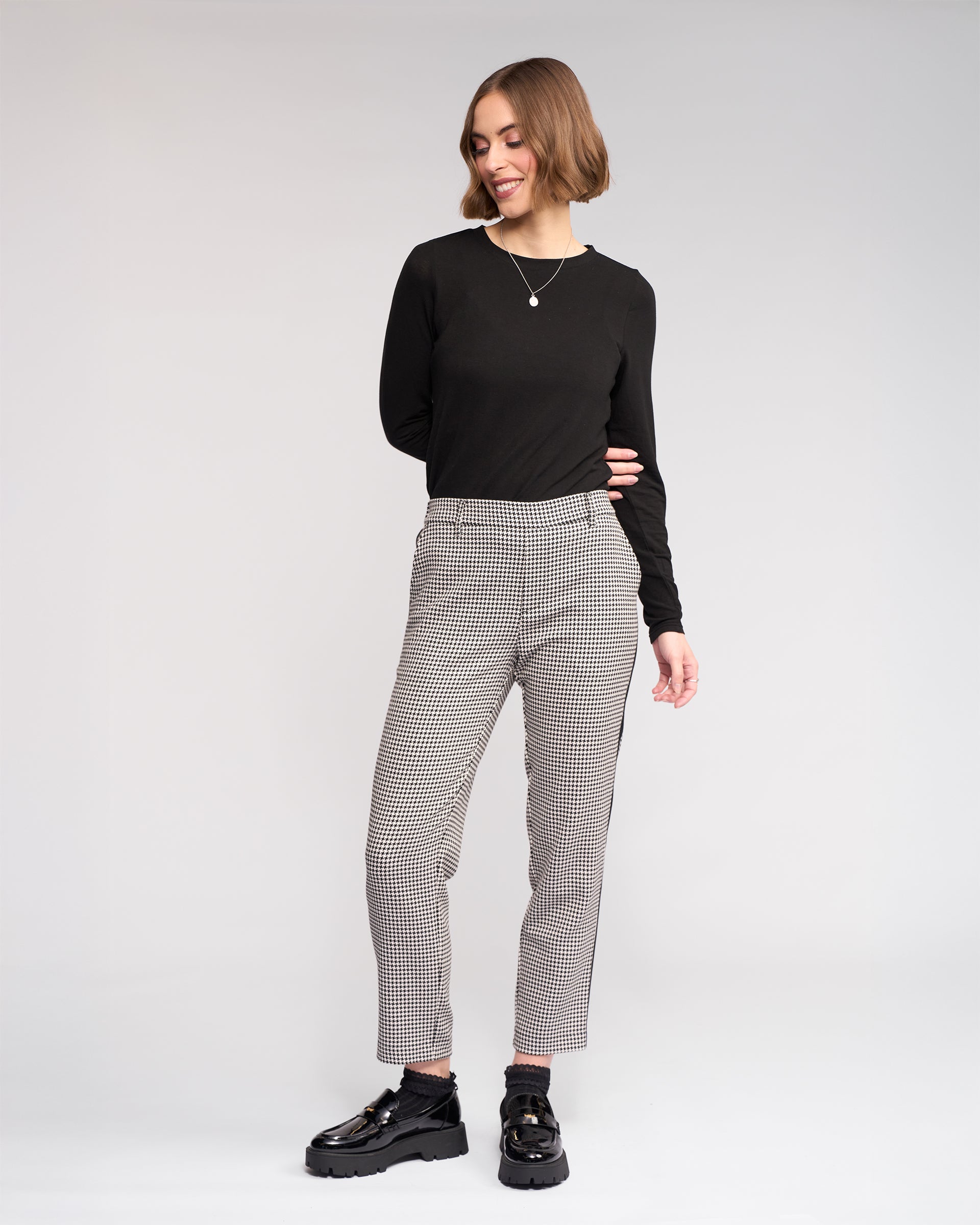 A woman stands smiling against a plain background, wearing the Knewe Label Como Pant - Summit paired with a black long-sleeve top and chunky shoes. She has short, straight hair, a necklace, and slim-leg pants.