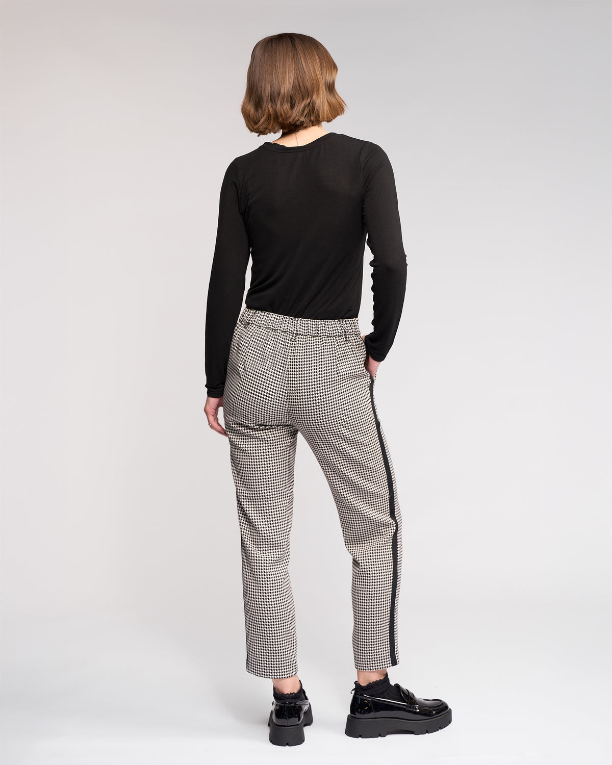 A person with short, wavy brown hair faces away, wearing a black top, Knewe Label's Como Pant - Summit with black side stripes, and black platform shoes against a plain light background.