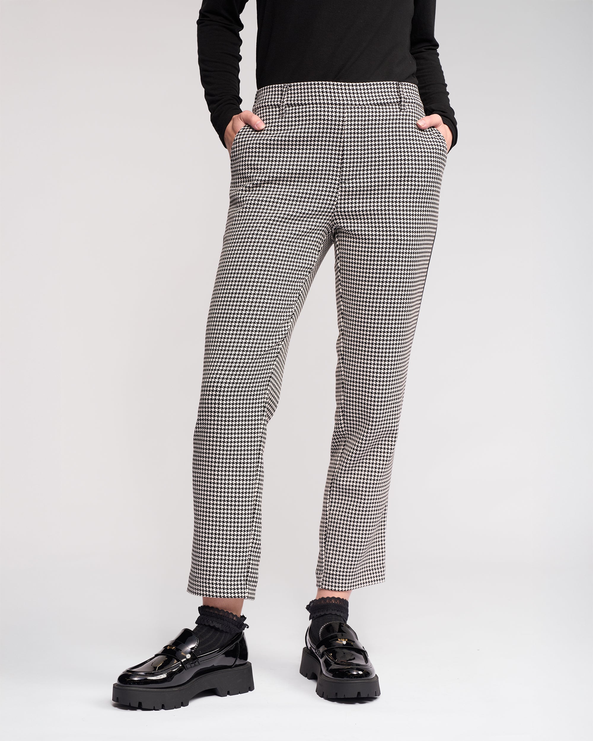 Wearing the Knewe Label Como Pant - Summit in black-and-white houndstooth, styled with a black long-sleeve top and chunky-soled shiny black loafers, hands in pockets, against a plain light background.