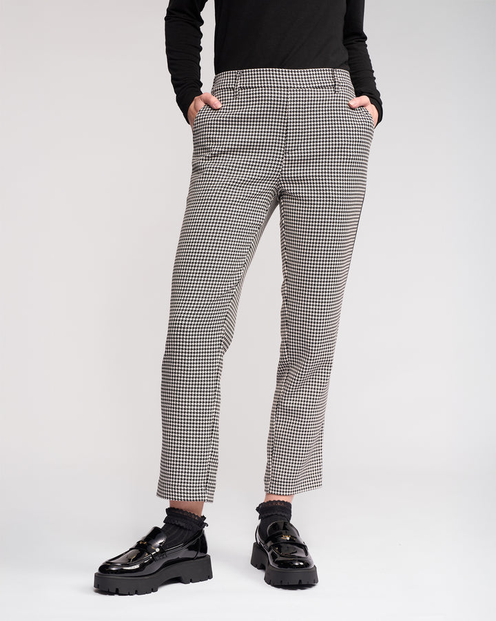 Wearing the Knewe Label Como Pant - Summit in black-and-white houndstooth, styled with a black long-sleeve top and chunky-soled shiny black loafers, hands in pockets, against a plain light background.