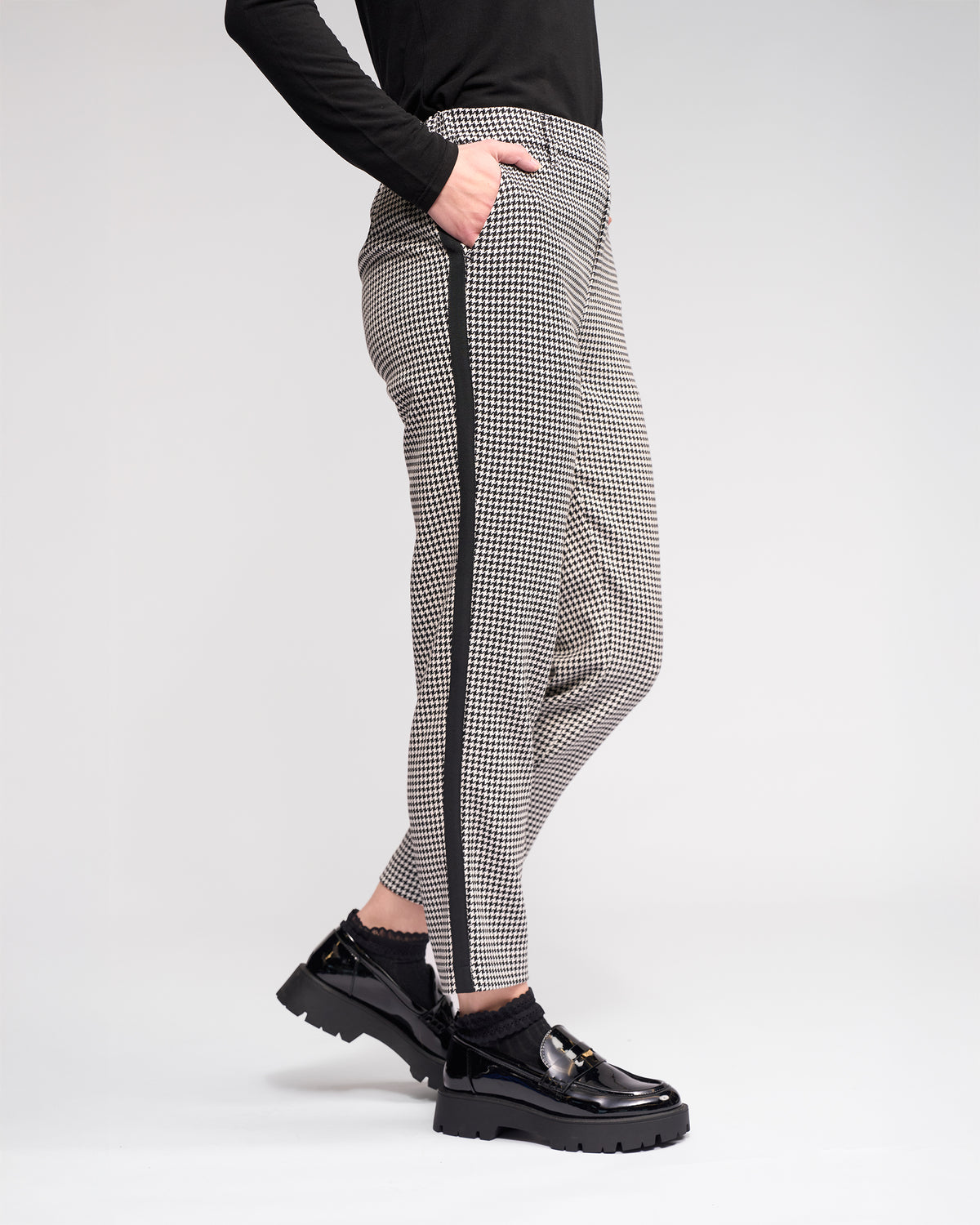 Someone wearing the Knewe Label Como Pant - Summit, featuring black-and-white houndstooth with a black side stripe, paired with black chunky loafers and a black long-sleeve top against a plain light background.