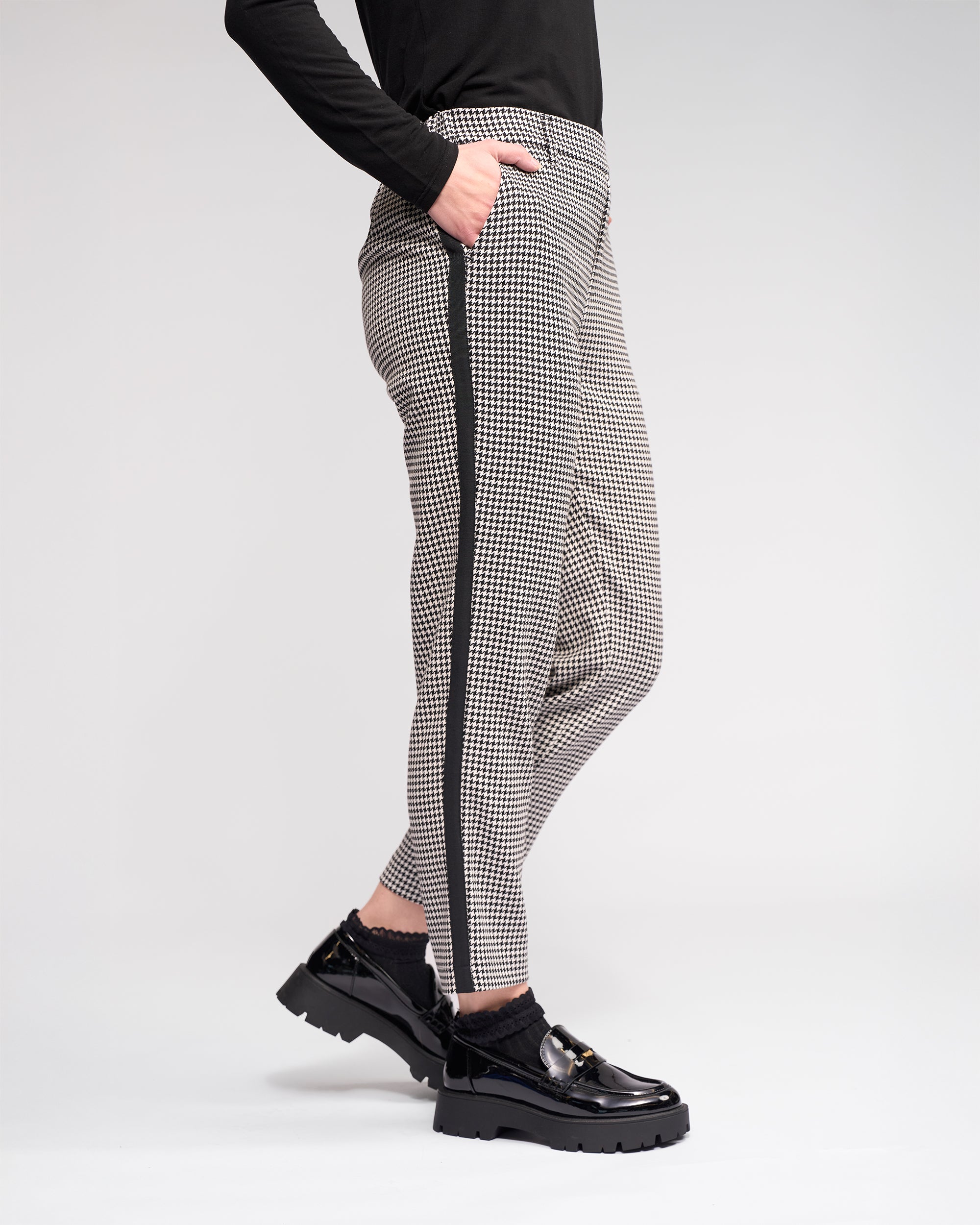 Someone wearing the Knewe Label Como Pant - Summit, featuring black-and-white houndstooth with a black side stripe, paired with black chunky loafers and a black long-sleeve top against a plain light background.