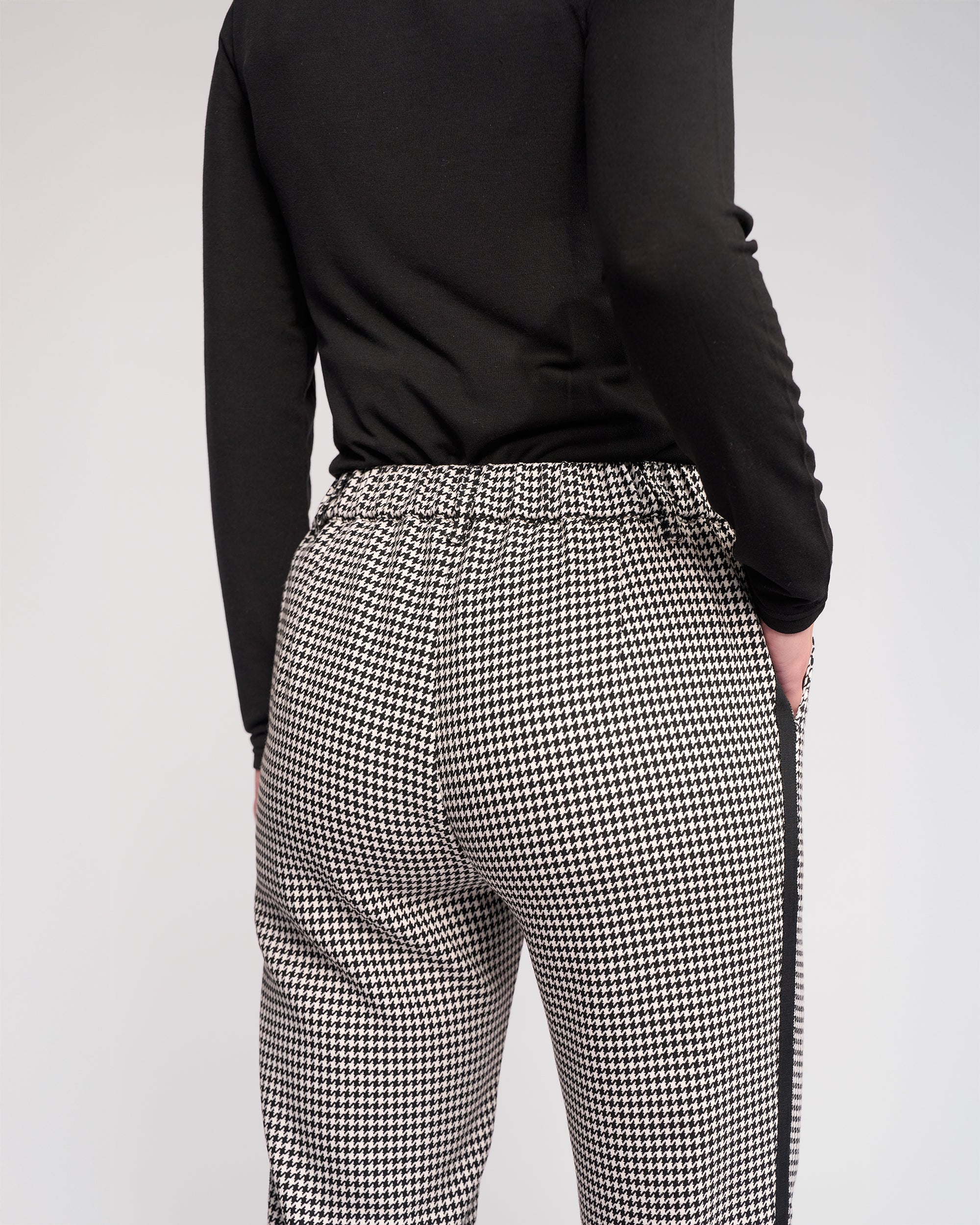 Shown from the back against a plain light background, a person wears a black long-sleeve top and the Knewe Label Como Pant – Summit: black-and-white houndstooth, slim-leg, ankle-grazer pants.