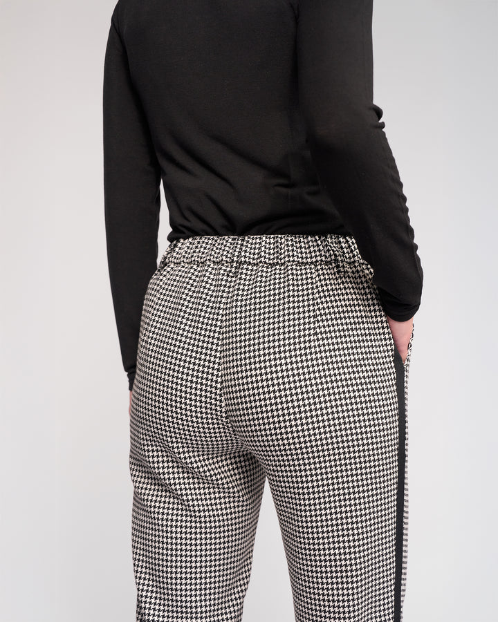Shown from the back against a plain light background, a person wears a black long-sleeve top and the Knewe Label Como Pant – Summit: black-and-white houndstooth, slim-leg, ankle-grazer pants.
