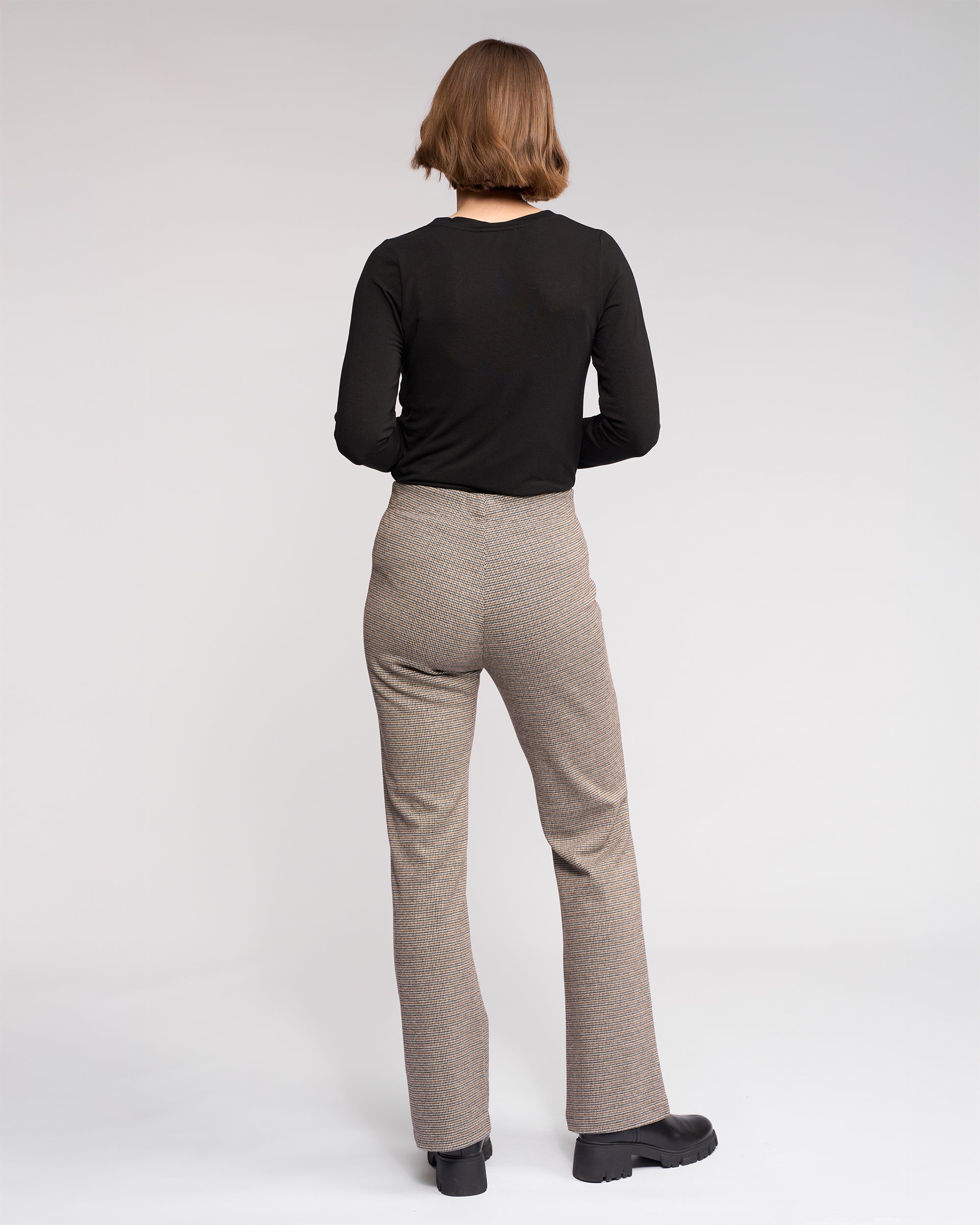 A person with short, light brown hair stands facing away, wearing a black long-sleeve top, Knewe Label's Como Flare Pant - Cedar HT in beige houndstooth stretch fabric, and black shoes against a plain light background.