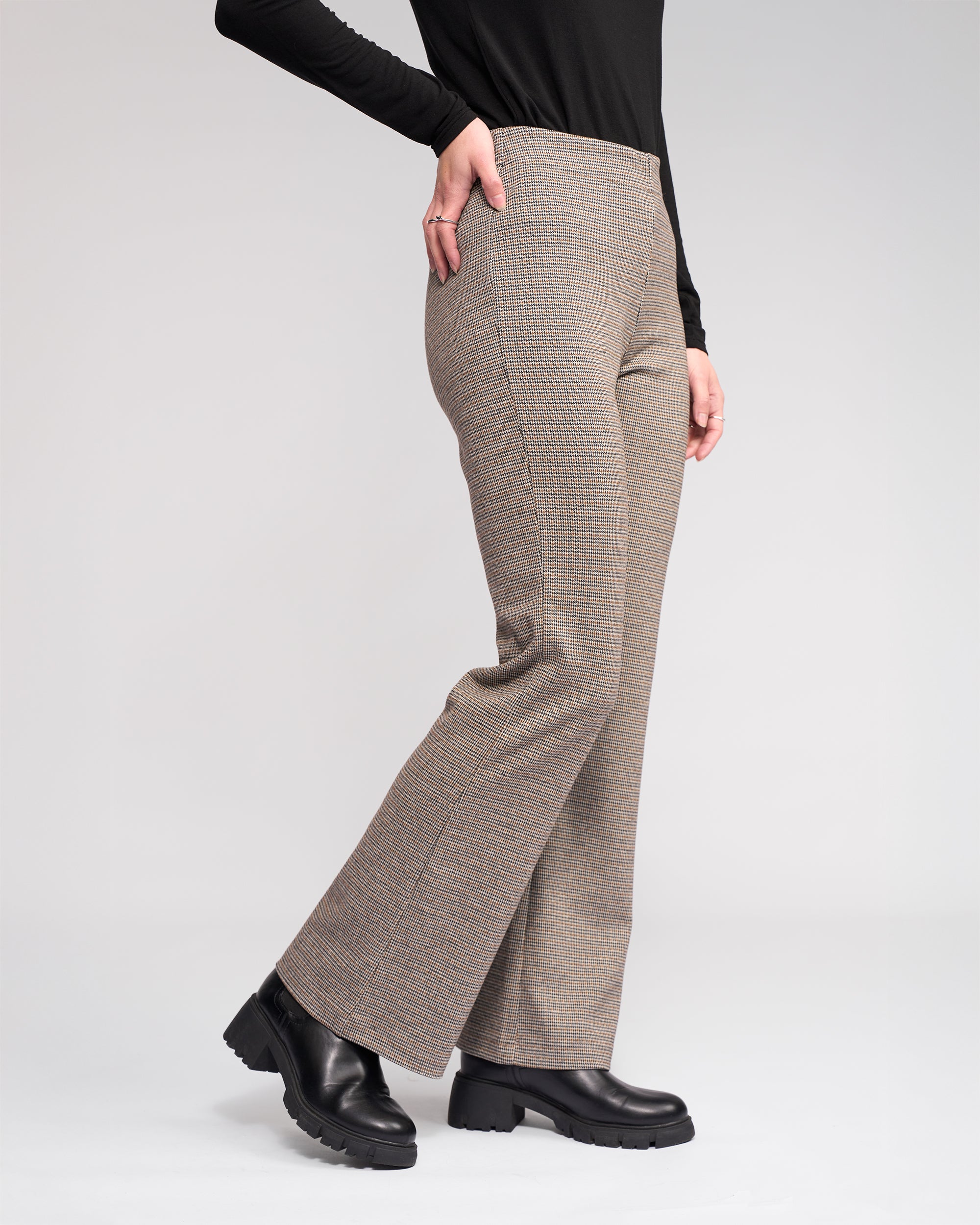 Wearing Knewe Label's Como Flare Pant - Cedar HT, a person poses in stylish high-waisted houndstooth pants with a black long-sleeve top and black chunky-heeled boots against a plain light background, hand on hip.