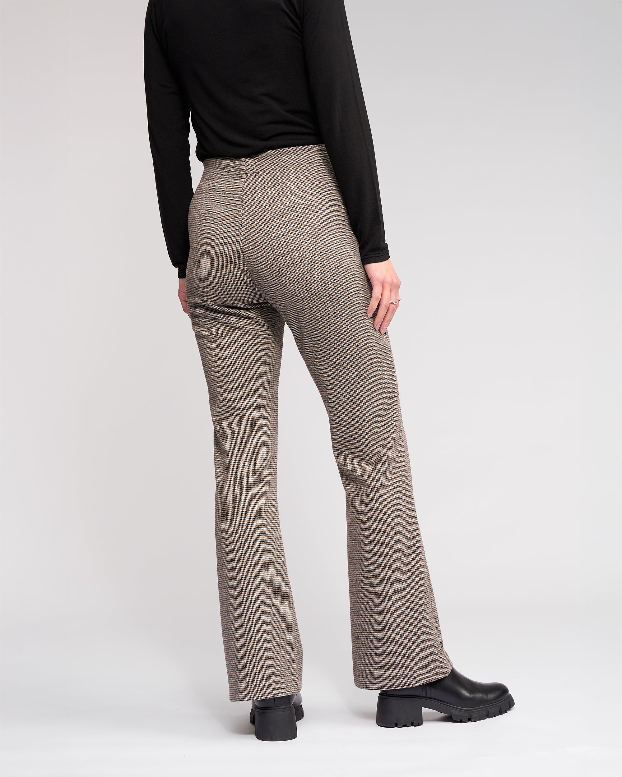 A person faces away, wearing a black long-sleeve top, black chunky-heeled boots, and Knewe Label's Como Flare Pant - Cedar HT in brown houndstooth stretch fabric, set against a plain light background.
