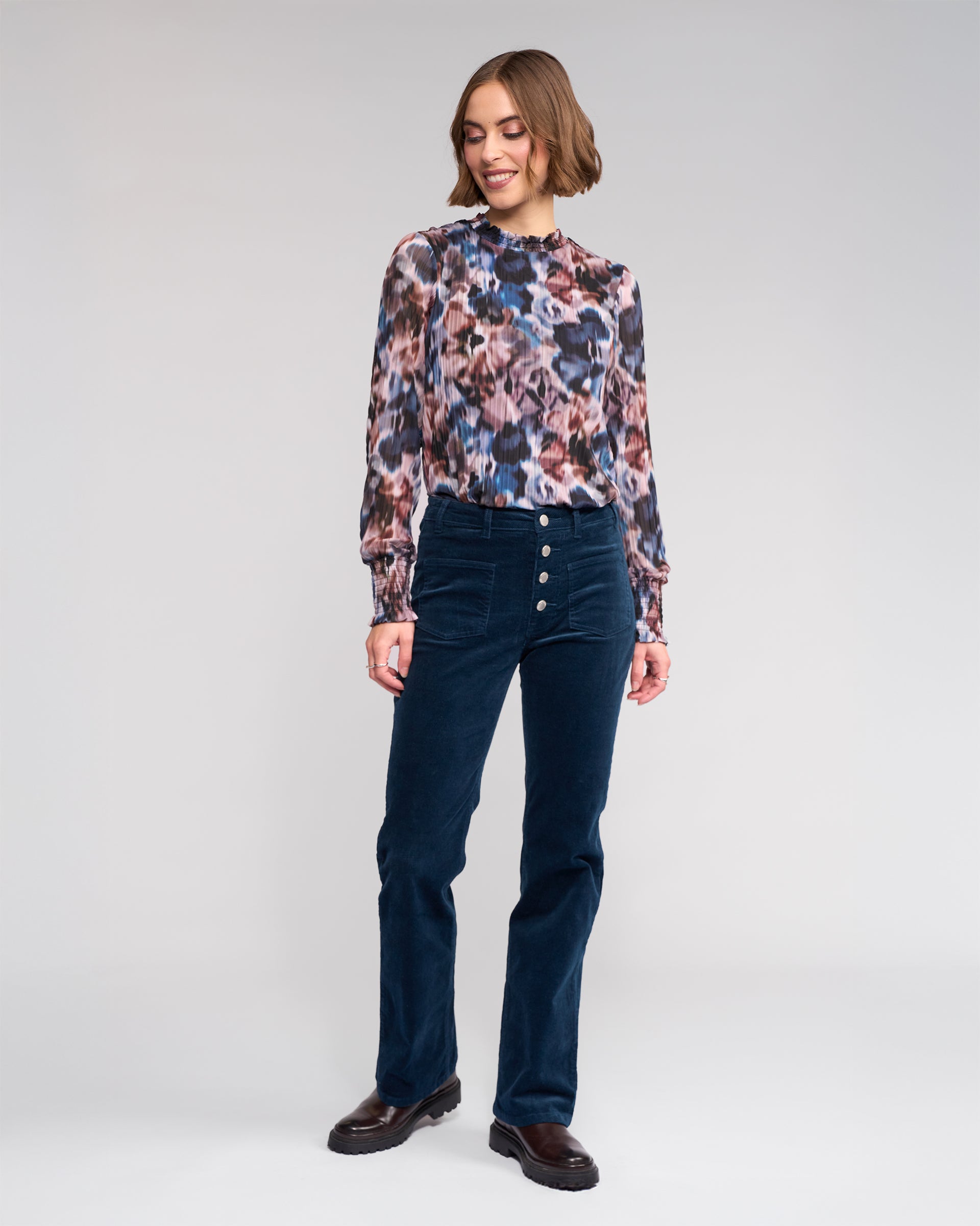 A woman with short hair smiles and looks to the side, wearing a long-sleeve multicolored floral blouse and Knewe Label's Potter Cord - Airforce bootcut corduroy pants against a plain background.