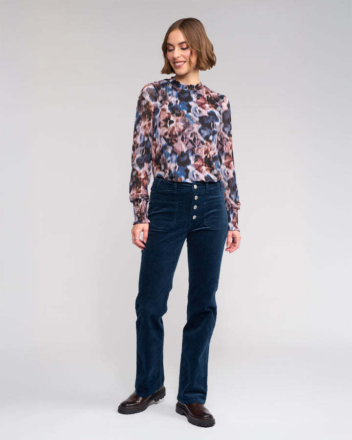 A woman with short hair smiles and looks to the side, wearing a long-sleeve multicolored floral blouse and Knewe Label's Potter Cord - Airforce bootcut corduroy pants against a plain background.