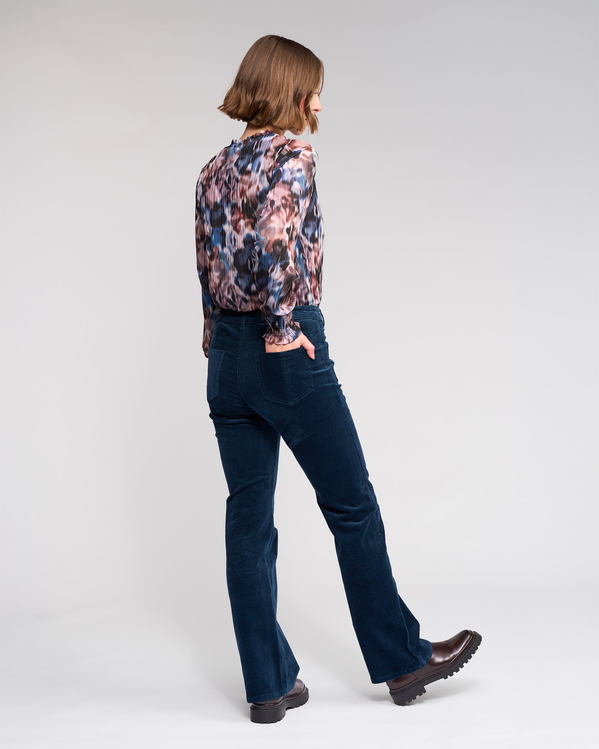 A person with shoulder-length brown hair, wearing a patterned blouse, Potter Cord - Airforce pants by Knewe Label, and brown shoes stands sideways against a plain light background.