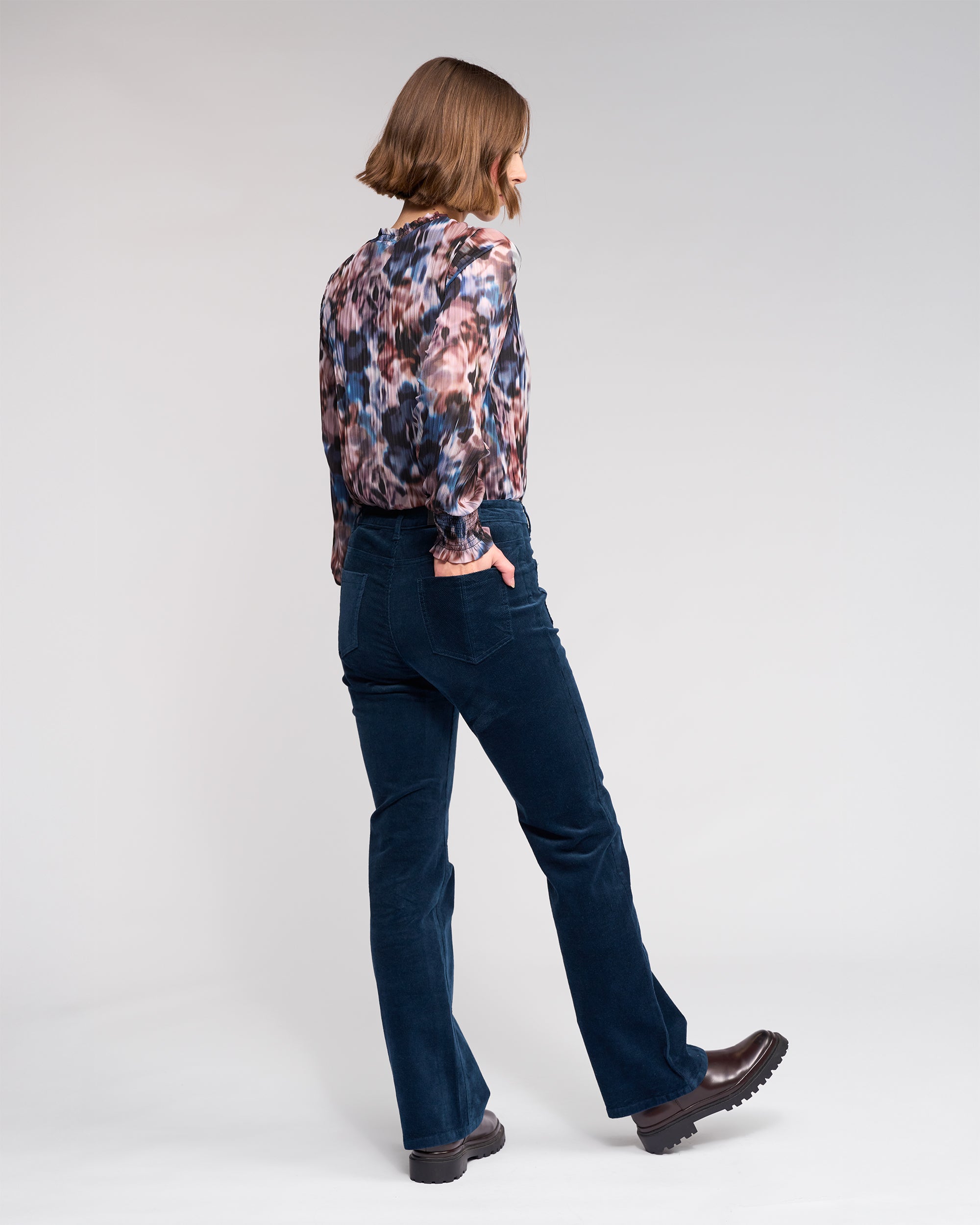 A person with shoulder-length brown hair, wearing a patterned blouse, Potter Cord - Airforce pants by Knewe Label, and brown shoes stands sideways against a plain light background.