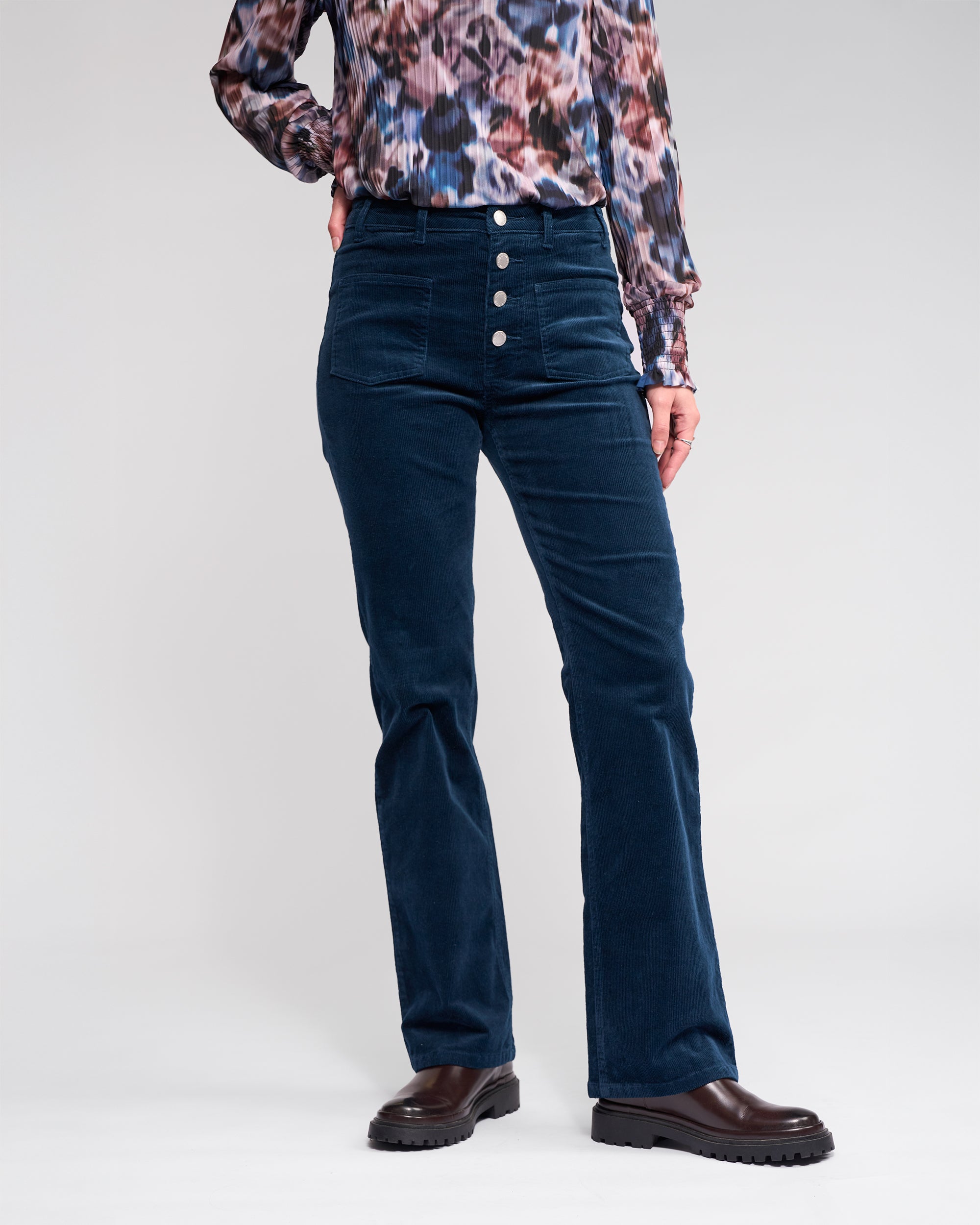 A person wears Knewe Label's Potter Cord - Airforce bootcut corduroy pants with visible buttons, a patterned long-sleeve blouse, and dark brown shoes, standing against a plain light background.