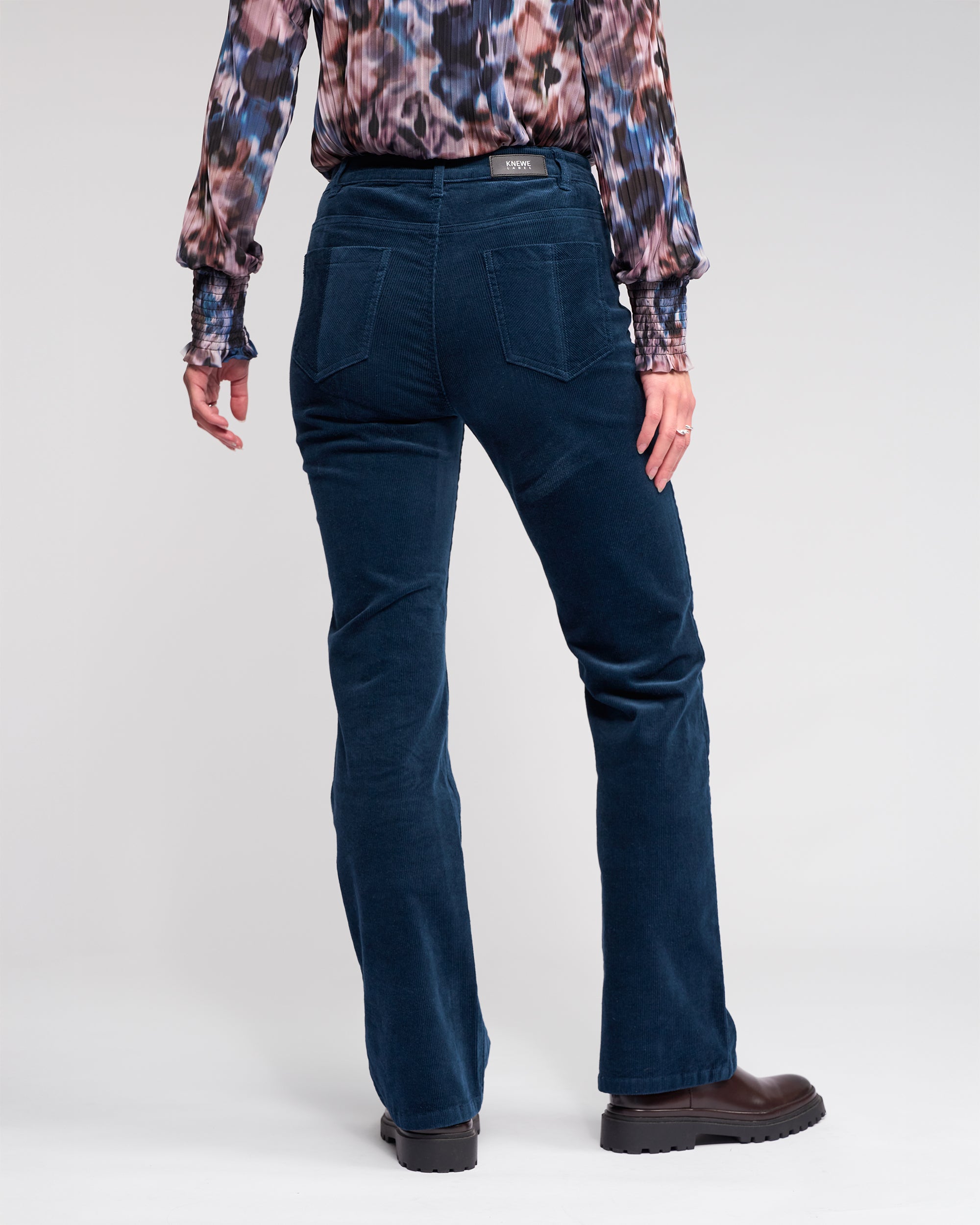 A person stands facing away against a plain light background, wearing Knewe Label's Potter Cord - Airforce bootcut corduroy pants, a patterned long-sleeve shirt, and black shoes.