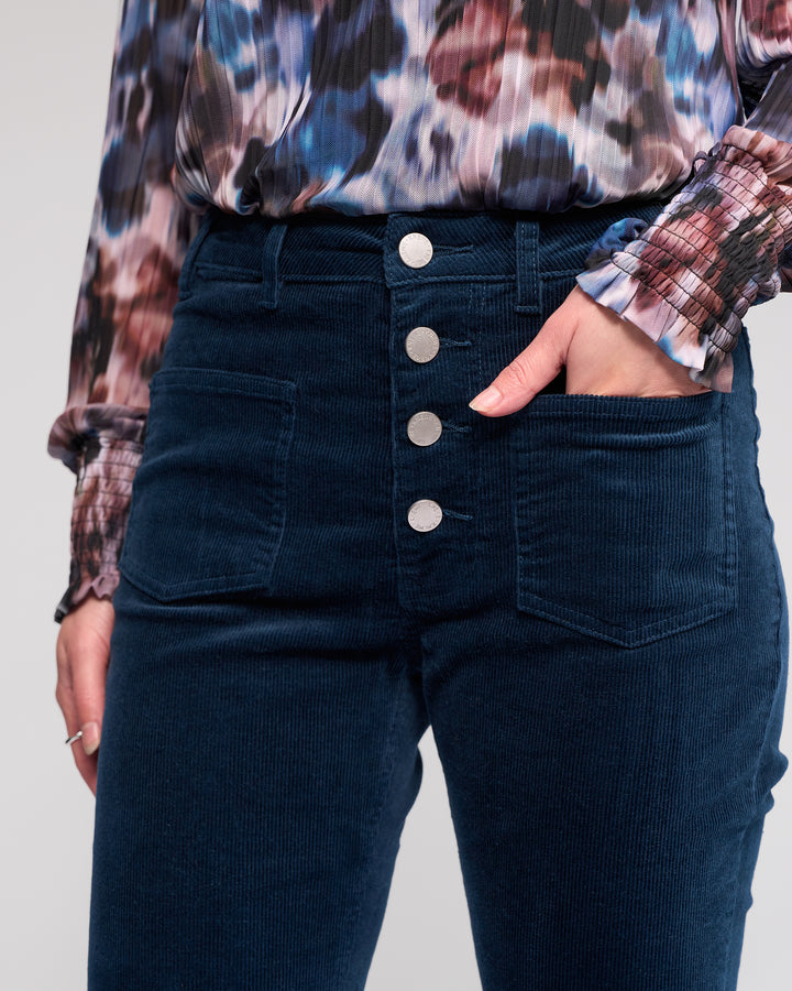 Wearing Knewe Label’s Potter Cord - Airforce wide-wale bootcut corduroy pants with four silver buttons and front pockets, paired with a colorful patterned long-sleeve blouse. One hand rests in a pocket.