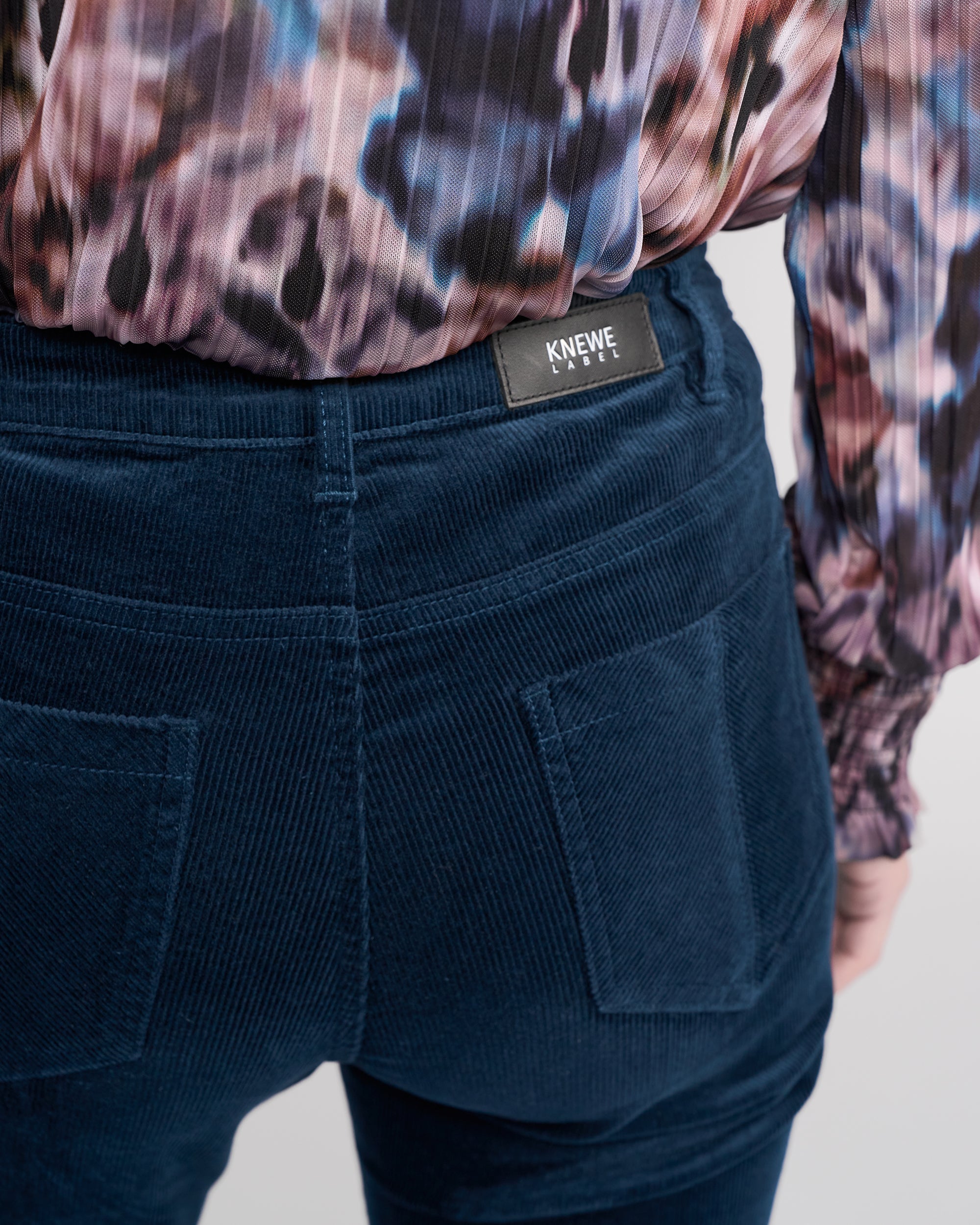 Close-up of a person wearing Knewe Label’s Potter Cord - Airforce: stretch bootcut corduroy pants with large back pockets and branded label, paired with a colorful patterned long-sleeve blouse.