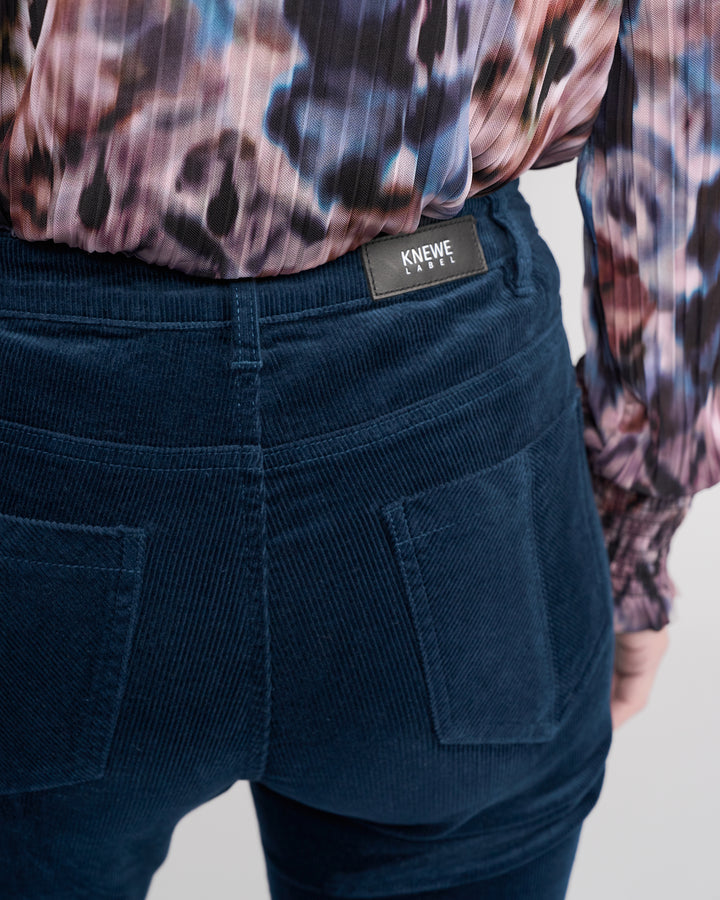 Close-up of a person wearing Knewe Label’s Potter Cord - Airforce: stretch bootcut corduroy pants with large back pockets and branded label, paired with a colorful patterned long-sleeve blouse.