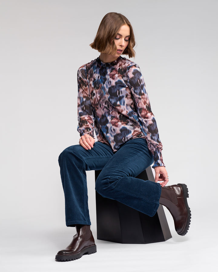 A woman with a bob haircut sits on a black stool, wearing the Knewe Label Potter Cord - Airforce blouse and matching bootcut cords with brown ankle boots, looking down as she adjusts her boot against a plain, light backdrop.