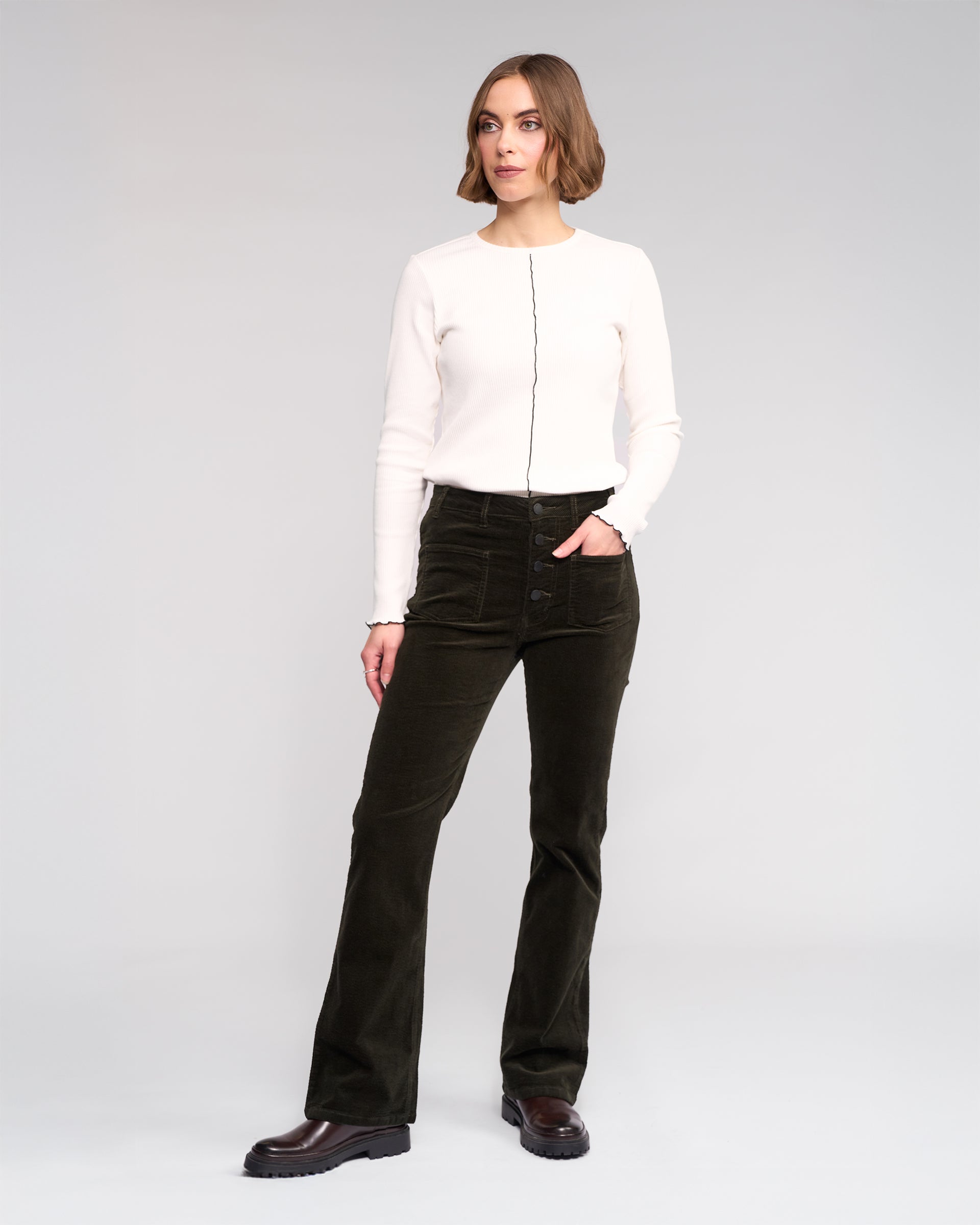 A woman with short hair poses confidently against a plain background wearing Knewe Label’s Potter Cord - Hunter dark green stretch wide-wale bootcut corduroy pants and a white long-sleeve top with a black vertical seam.