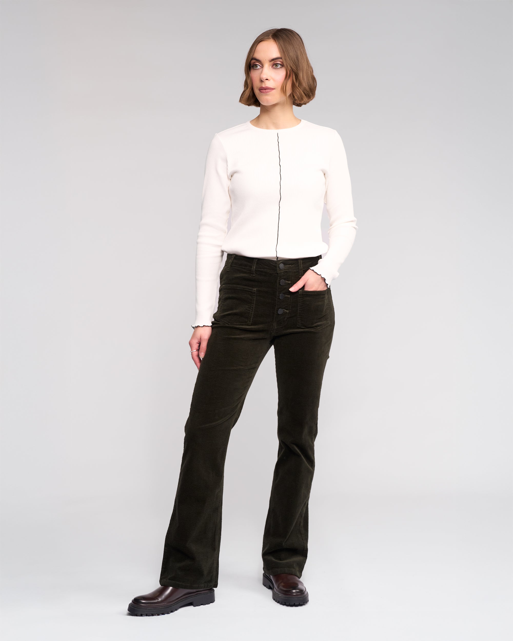 A woman with short hair poses confidently against a plain background wearing Knewe Label’s Potter Cord - Hunter dark green stretch wide-wale bootcut corduroy pants and a white long-sleeve top with a black vertical seam.