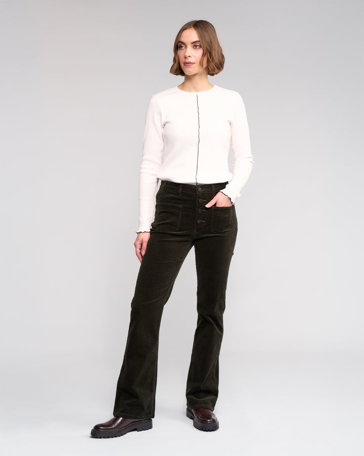 A woman with short hair poses confidently against a plain background wearing Knewe Label’s Potter Cord - Hunter dark green stretch wide-wale bootcut corduroy pants and a white long-sleeve top with a black vertical seam.