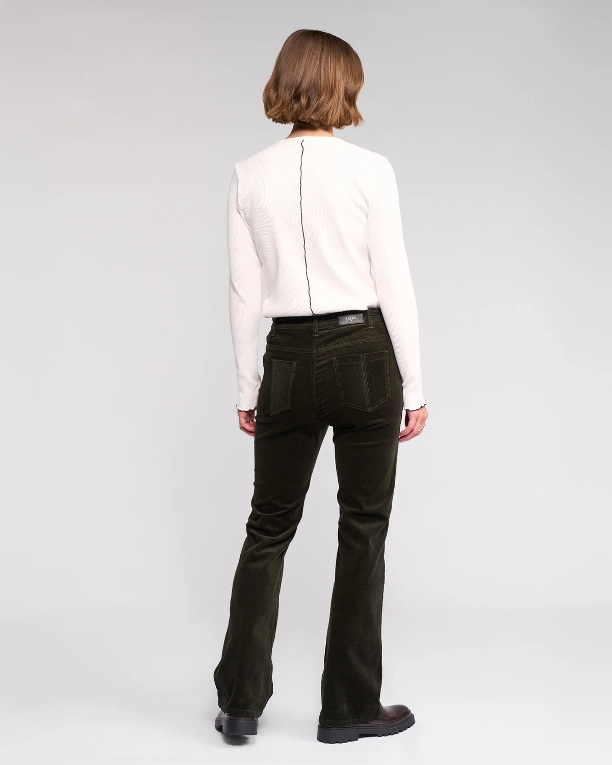 A person with short, light brown hair faces away, wearing a long-sleeve white top and Knewe Label's Potter Cord - Hunter wide-wale dark green bootcut corduroy pants, paired with black shoes against a plain light background.