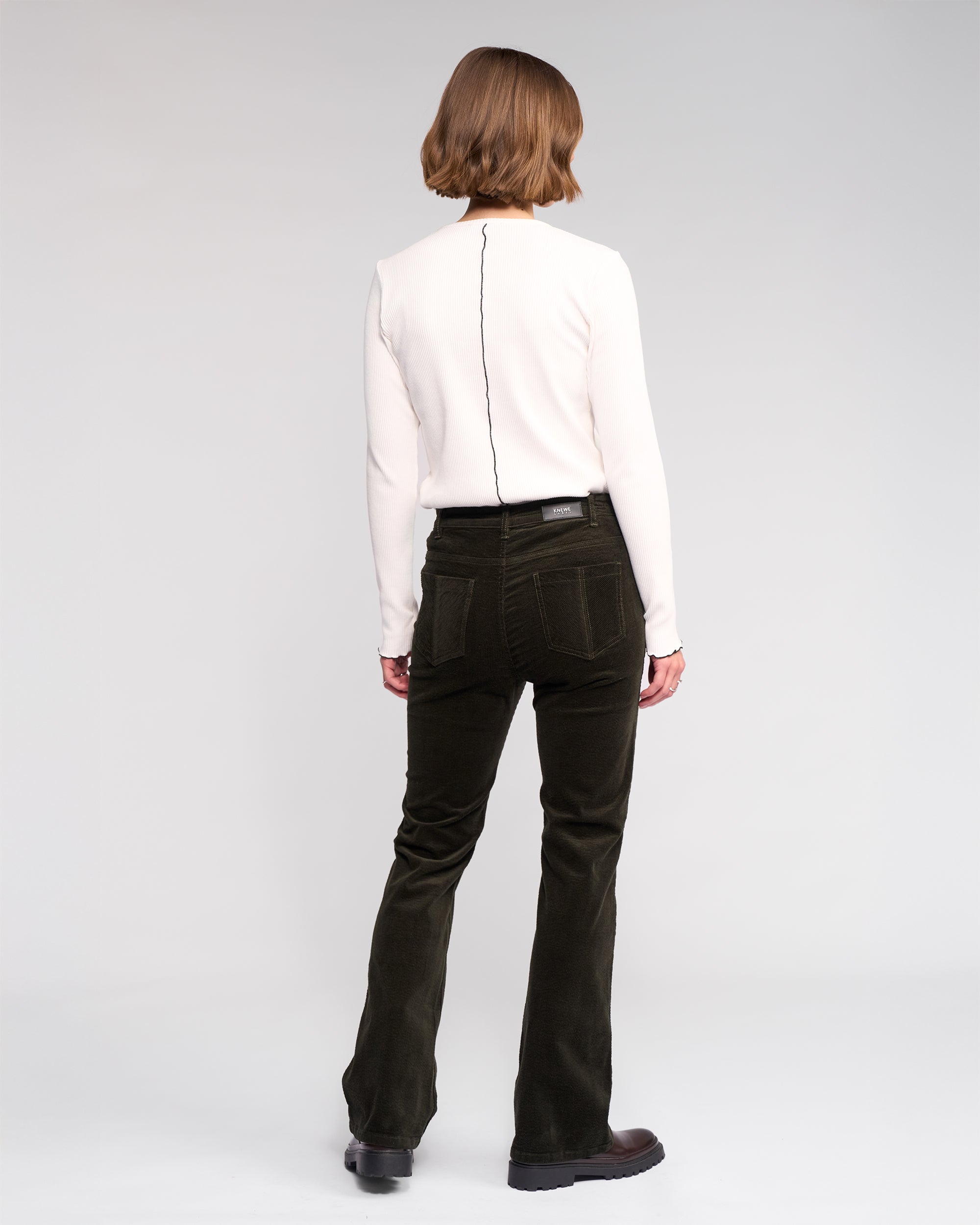 A person with short, light brown hair faces away, wearing a long-sleeve white top and Knewe Label's Potter Cord - Hunter wide-wale dark green bootcut corduroy pants, paired with black shoes against a plain light background.