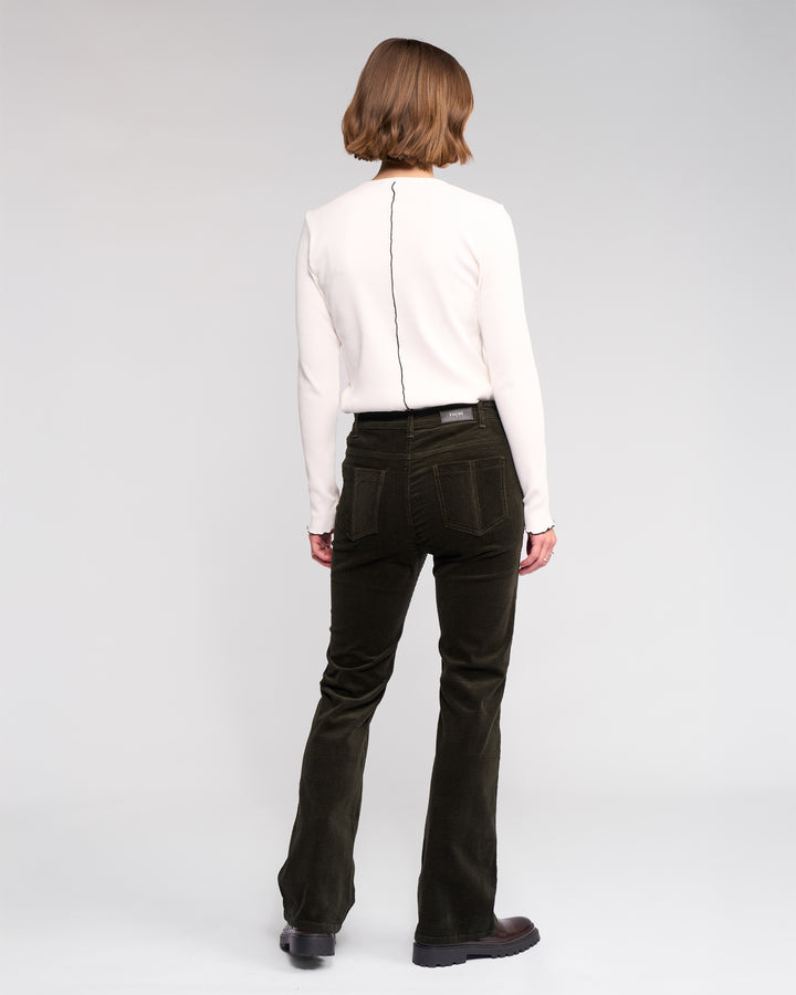 A person with short, light brown hair faces away, wearing a long-sleeve white top and Knewe Label's Potter Cord - Hunter wide-wale dark green bootcut corduroy pants, paired with black shoes against a plain light background.
