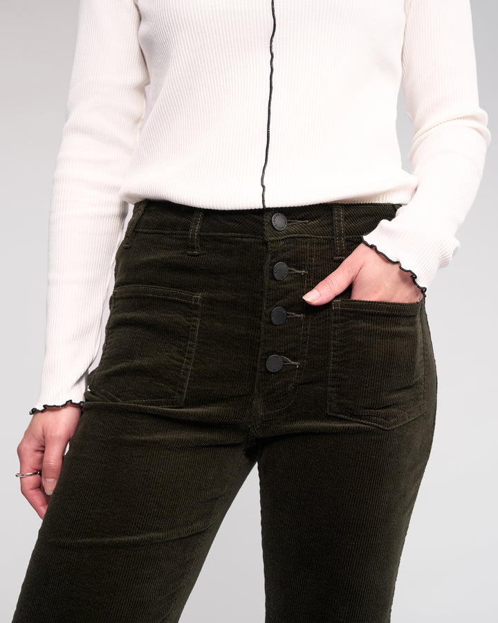 A person wears Knewe Label's Potter Cord - Hunter dark green bootcut corduroy pants, made from stretch wide-wale cord, paired with a white long-sleeve top with black trim, standing with one hand in their pocket.