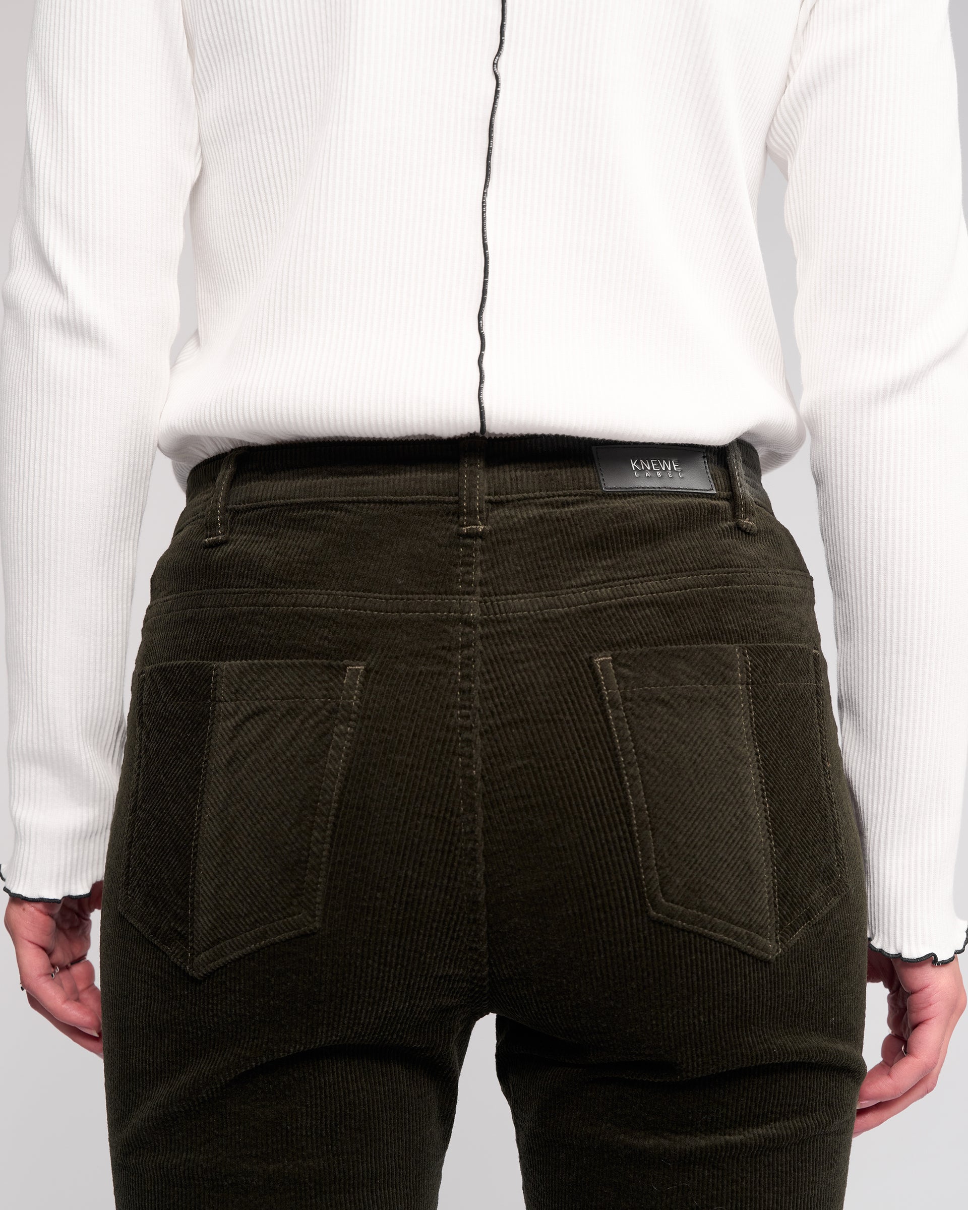 A person is seen from the back wearing a ribbed white long-sleeve top and Knewe Label Potter Cord - Hunter dark green wide-wale bootcut corduroy pants, with hands resting at their sides.