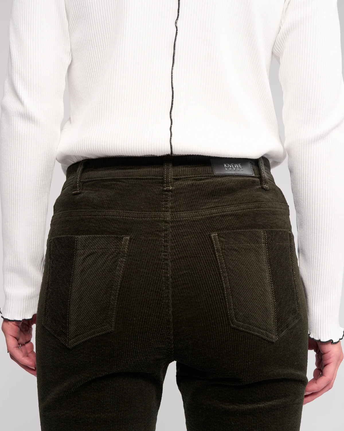 A person is seen from the back wearing a ribbed white long-sleeve top and Knewe Label Potter Cord - Hunter dark green wide-wale bootcut corduroy pants, with hands resting at their sides.