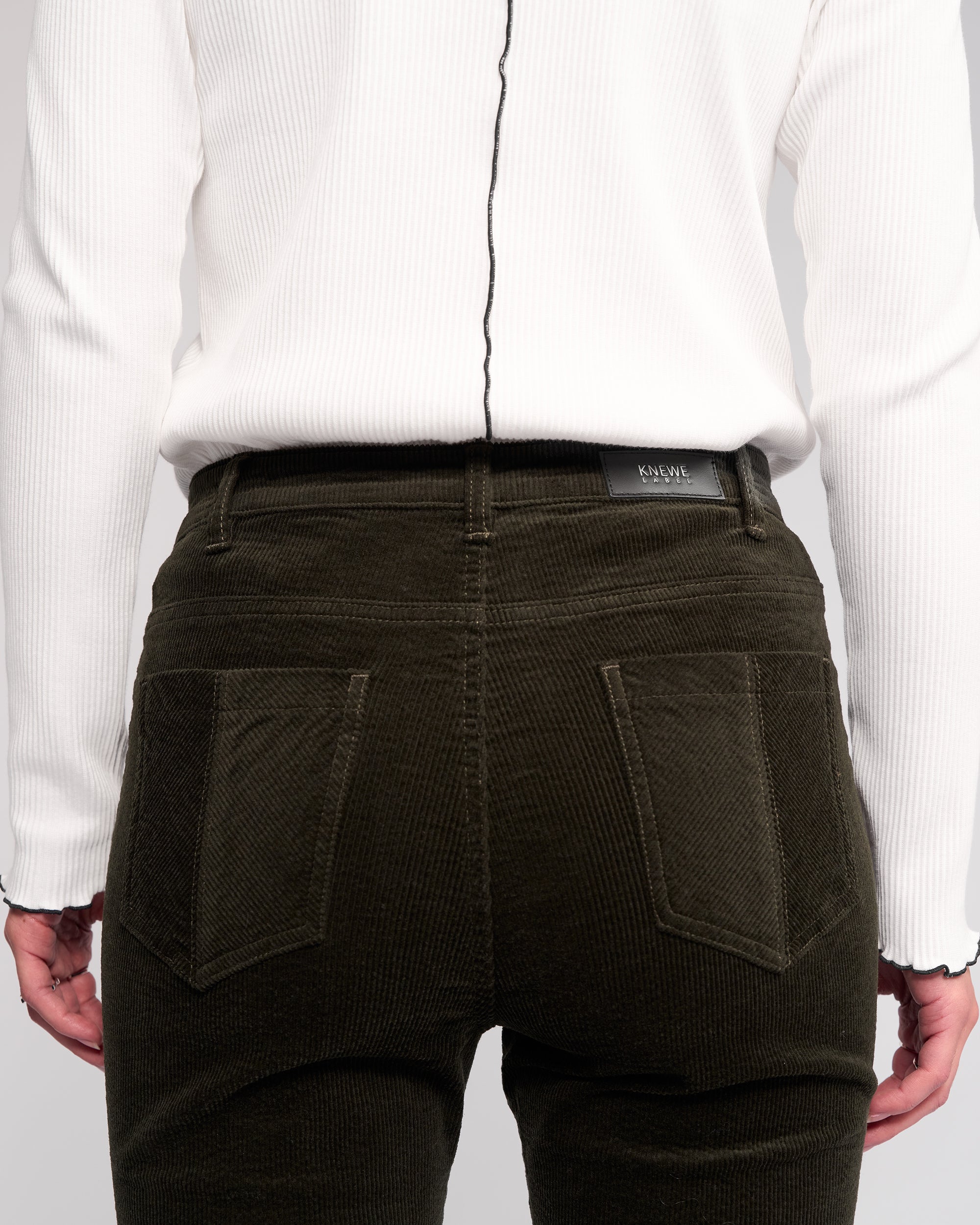 A person is seen from the back wearing a ribbed white long-sleeve top and Knewe Label Potter Cord - Hunter dark green wide-wale bootcut corduroy pants, with hands resting at their sides.